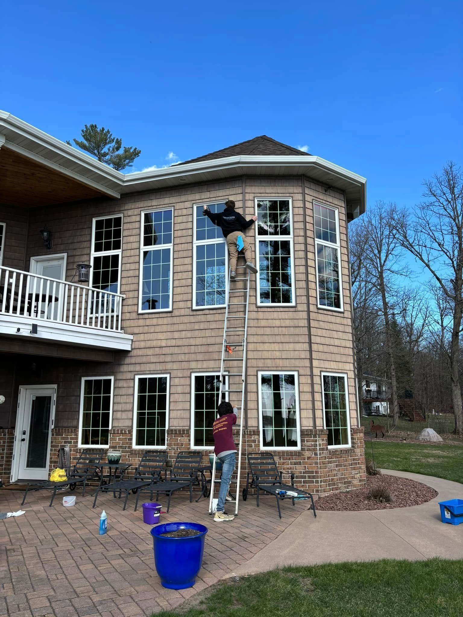 A person stands on a tall ladder cleaning the windows of a multi-story brick house on a sunny day.