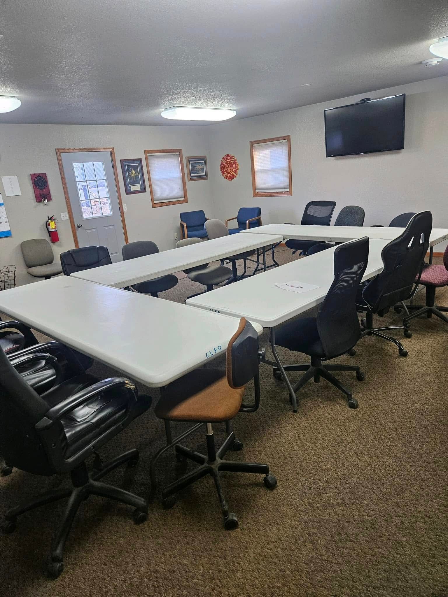 A conference room with U-shaped tables, several black office chairs, a TV on the wall, and windows in a light-colored room.