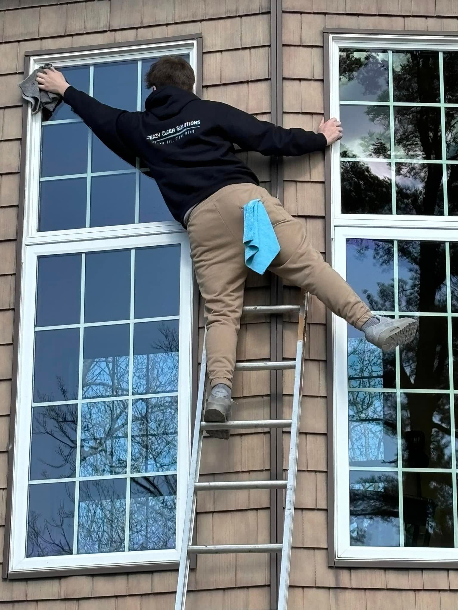 A person on a ladder balances between two windows while cleaning them with a cloth.