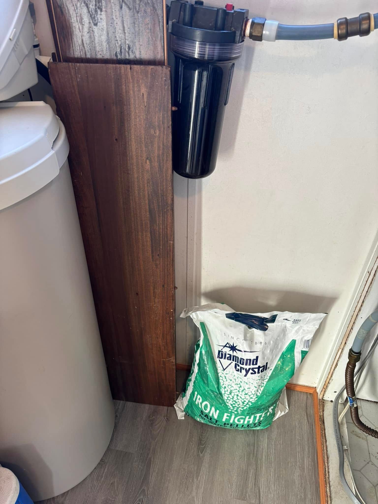A water softener tank and a wall-mounted water filter next to a bag of Iron Fighter water softener salt on a floor.