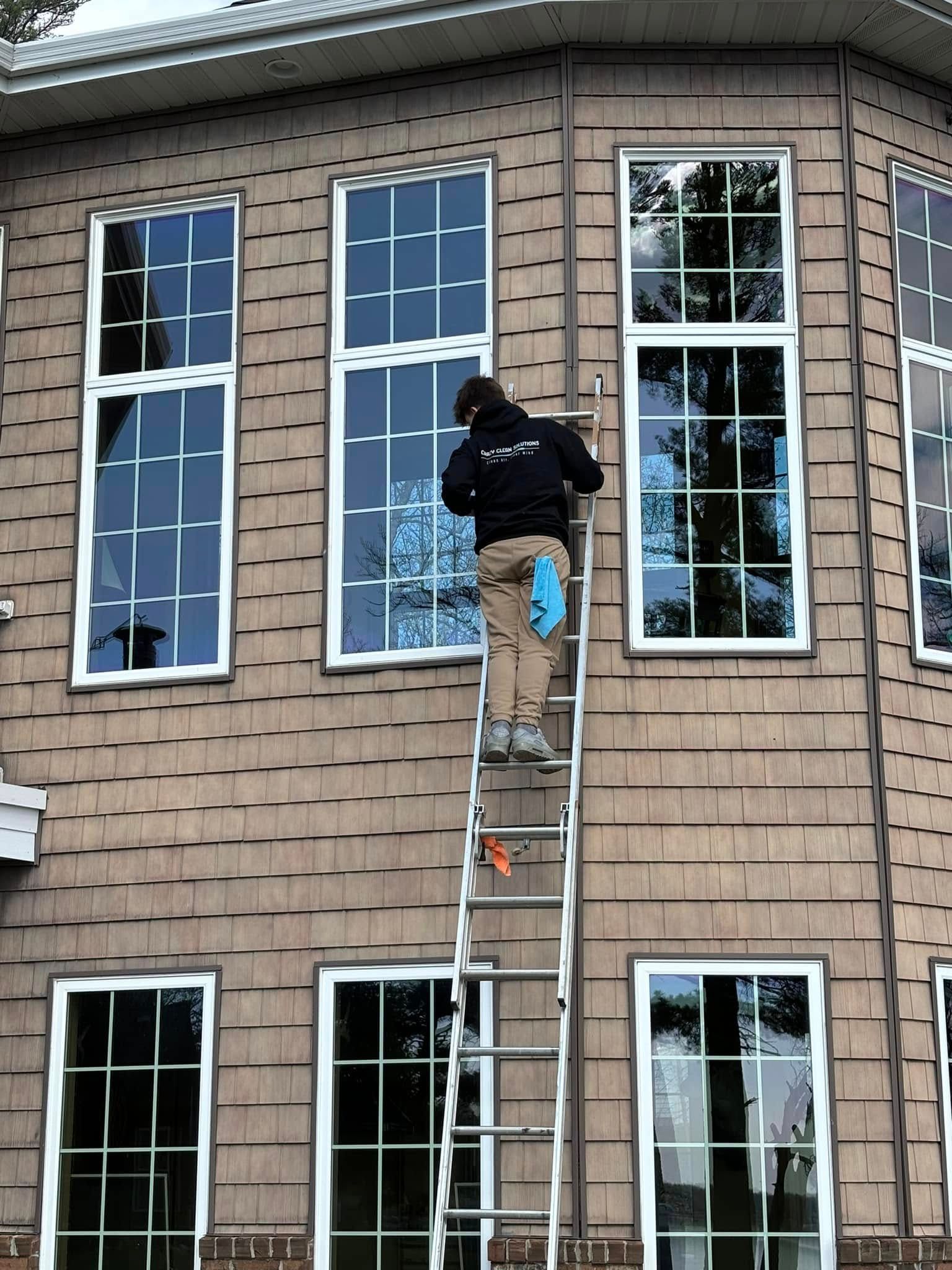 Person cleaning a window, standing on a ladder next to a brown shingled house with white-framed windows.
