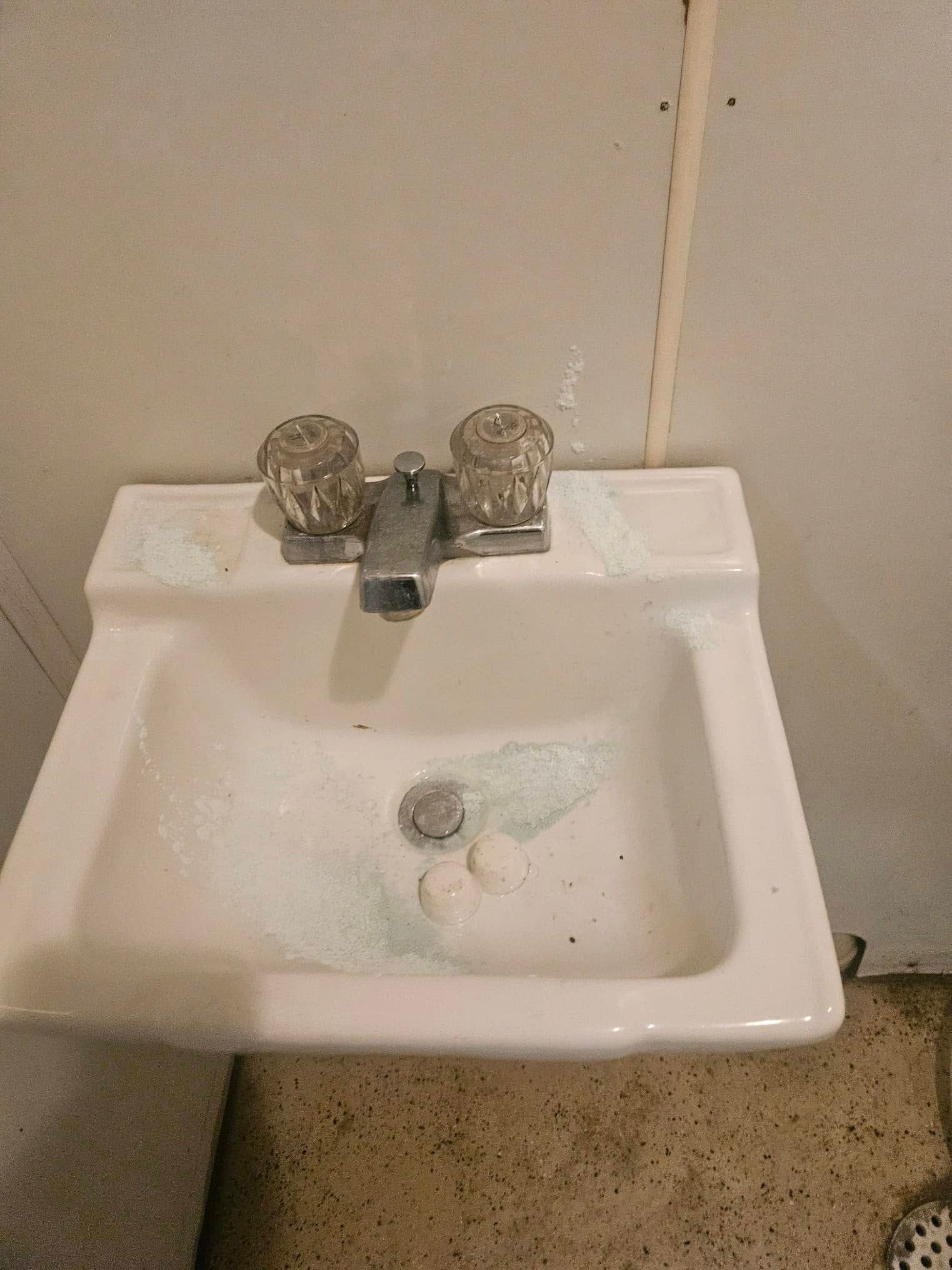 A wall-mounted, white porcelain bathroom sink with two clear acrylic knobs and a faucet, featuring residue in the basin.
