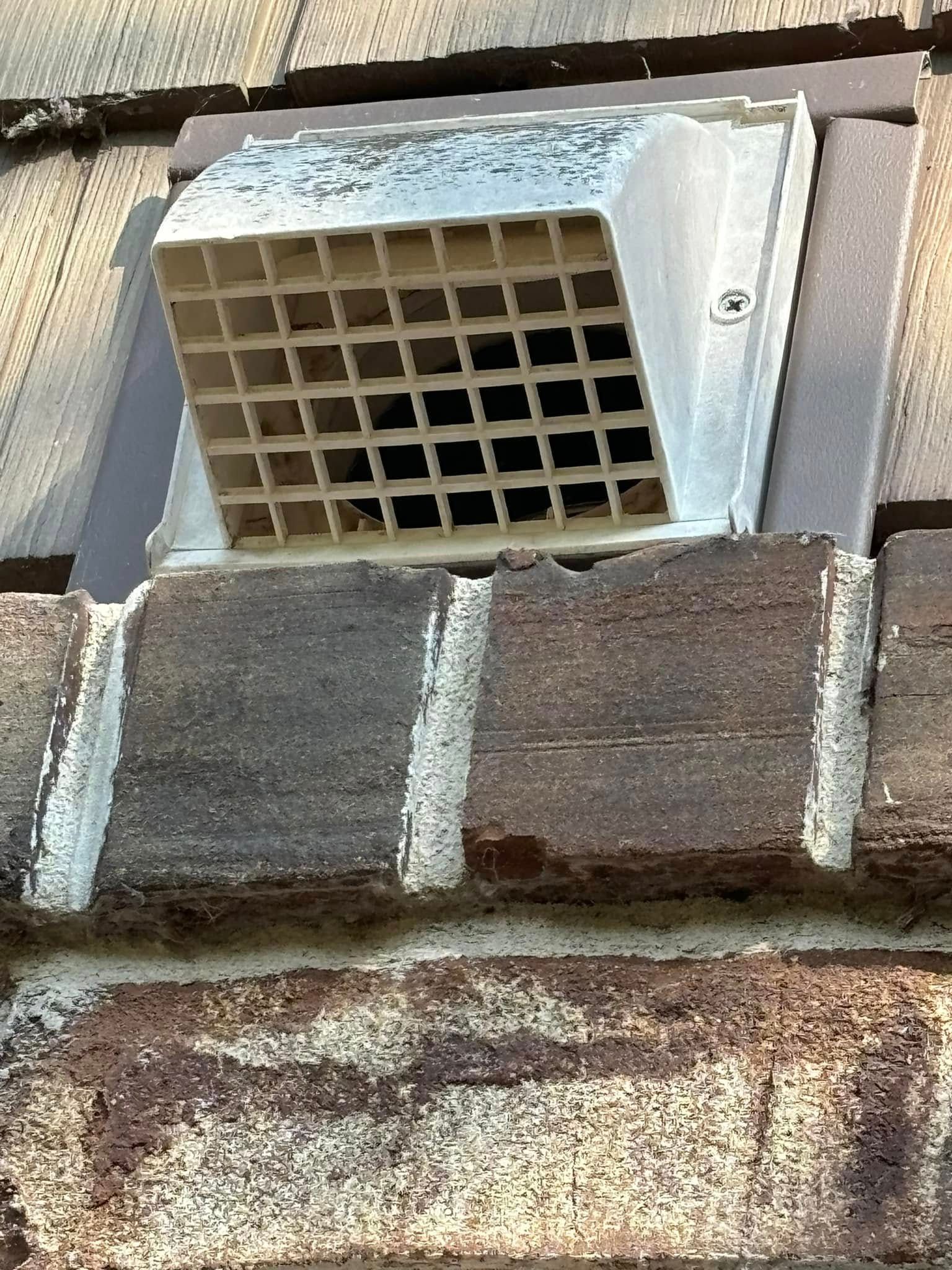 A beige, square-grid exhaust vent cover mounted on a brick exterior wall.
