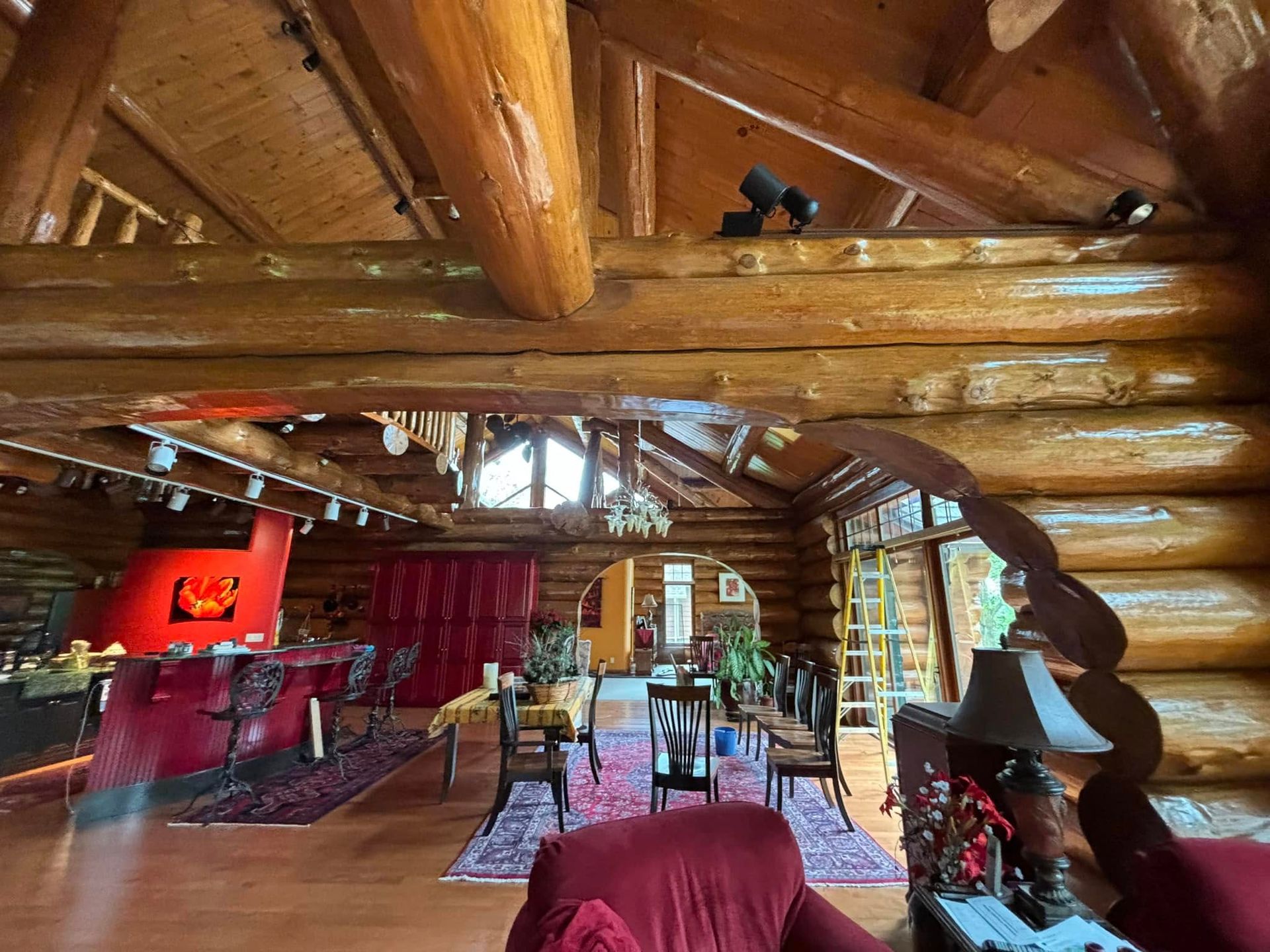 A spacious log cabin living area featuring exposed timber beams, a red kitchen island, and a dining set on a patterned rug.