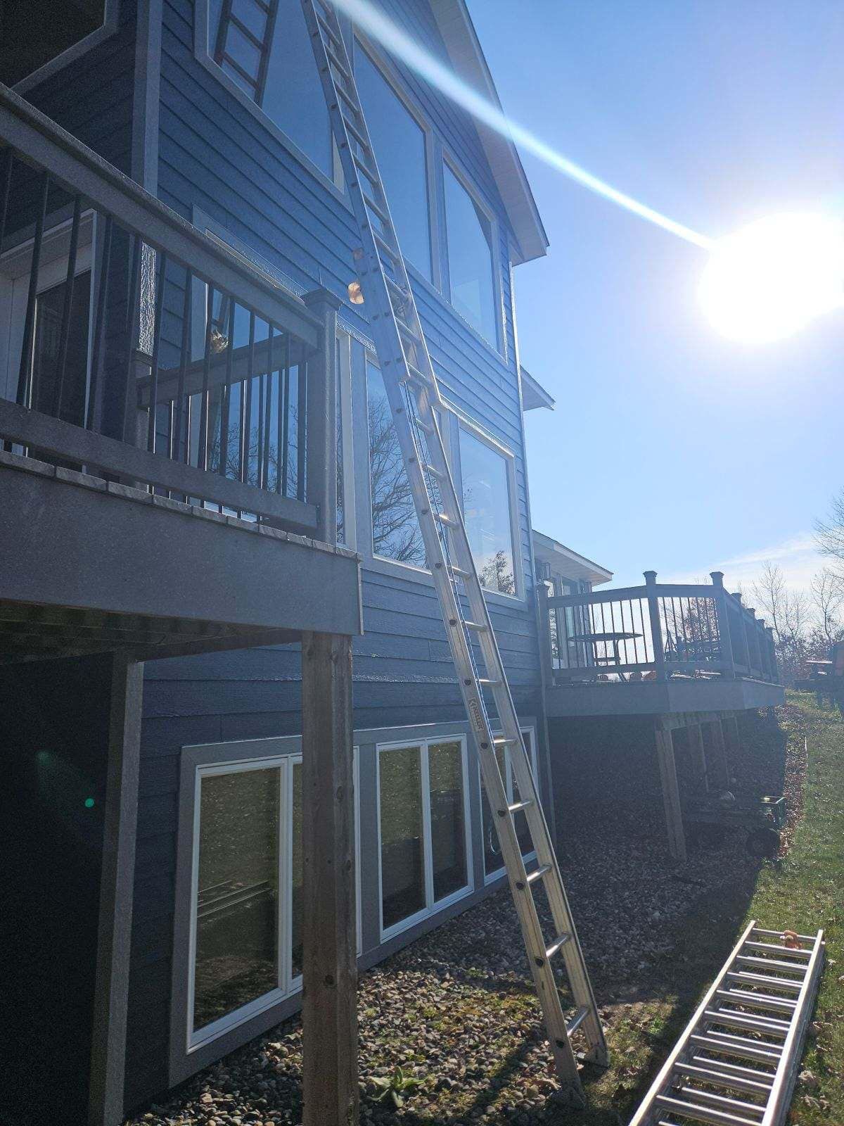 A tall extension ladder leans against the side of a blue-sided house near two decks, with a second ladder on the ground.