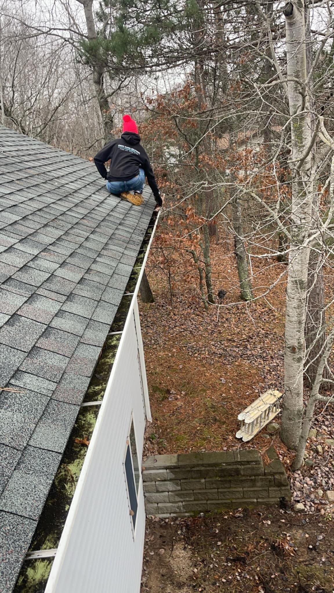 A person wearing a bright red hat and dark clothing sits on the edge of a sloped, shingled roof cleaning a gutter.