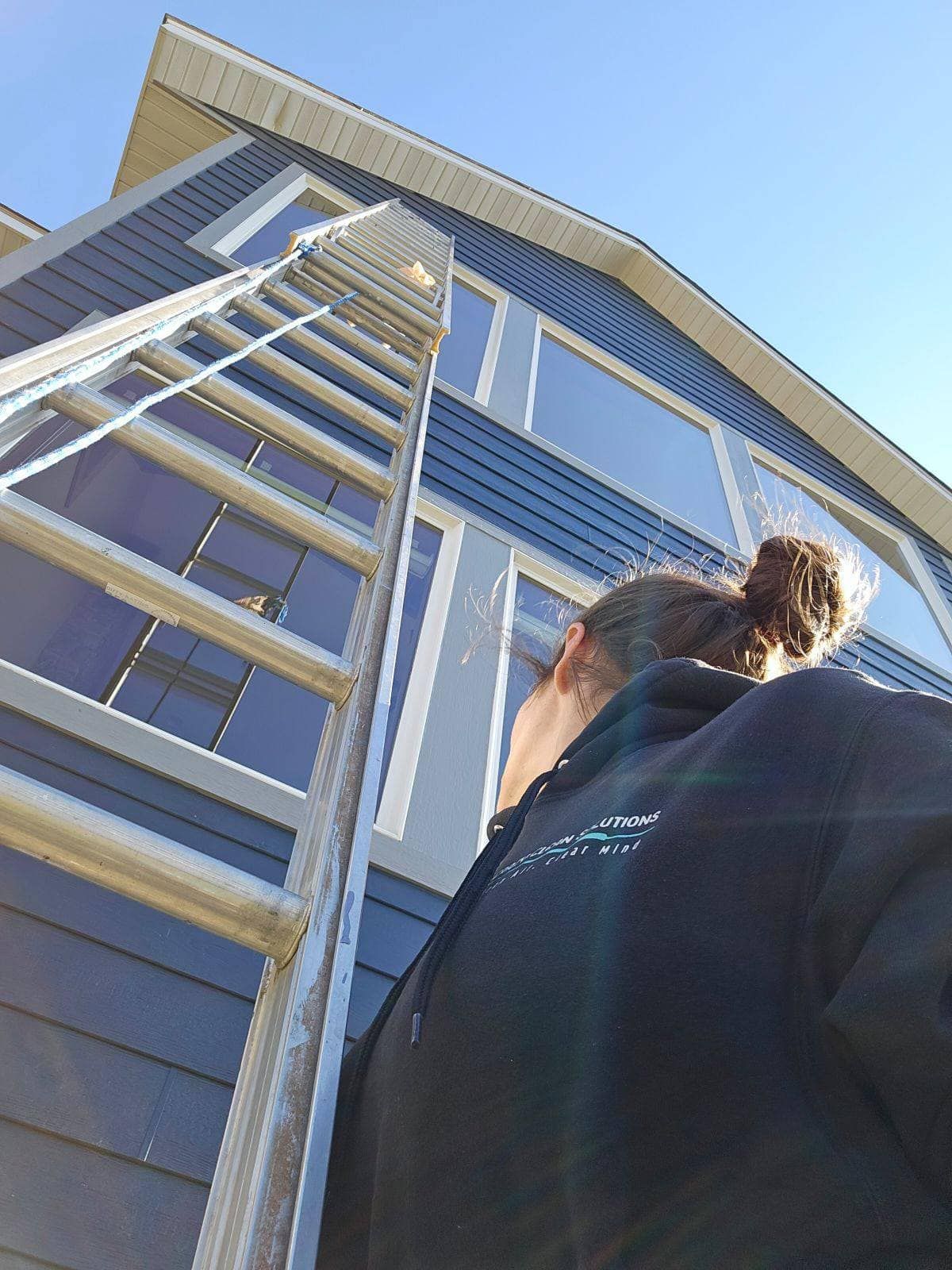 A person wearing a dark hoodie looks up at a tall ladder extending toward the top of a blue-sided house.