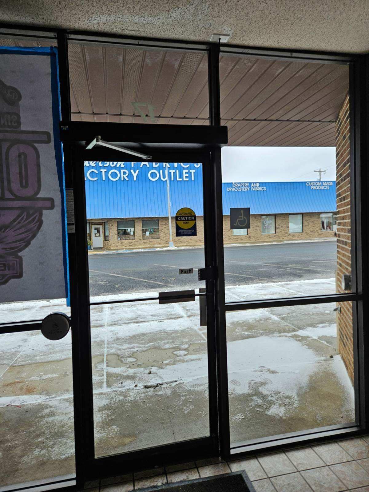 View through a glass storefront door looking out at a snowy parking lot and a building with a blue metal roof.