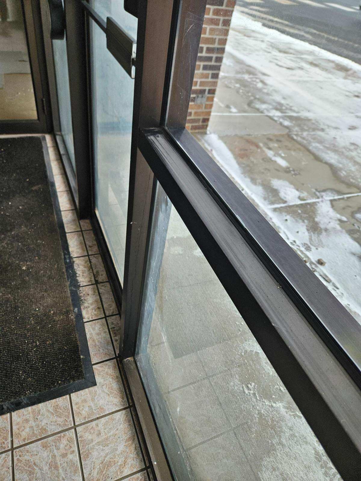 A view from inside looking out through a dark-framed glass door at a snowy sidewalk and a brick building wall.