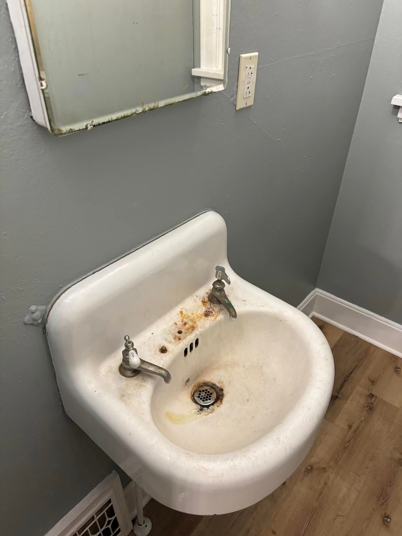 Wall-mounted white ceramic bathroom sink with two metal faucets, visible stains, and a mirror against grey walls.