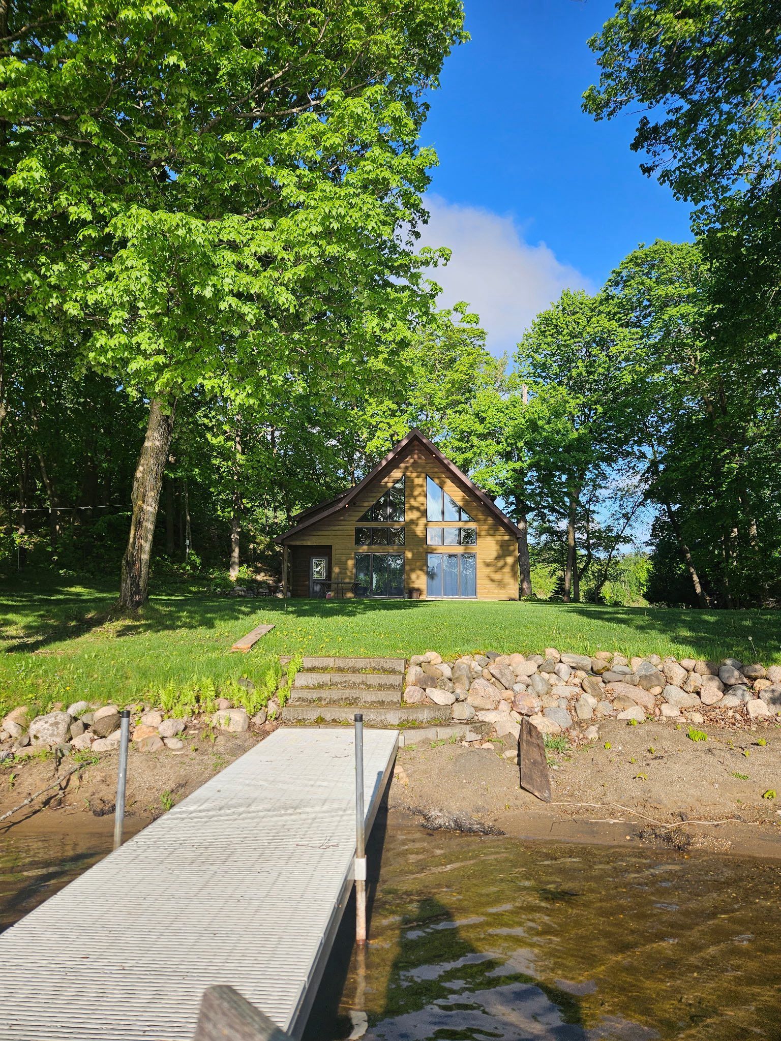 A modern A-frame cabin sits nestled in a lush, green forest on a grassy hill overlooking a wooden dock on a lake.