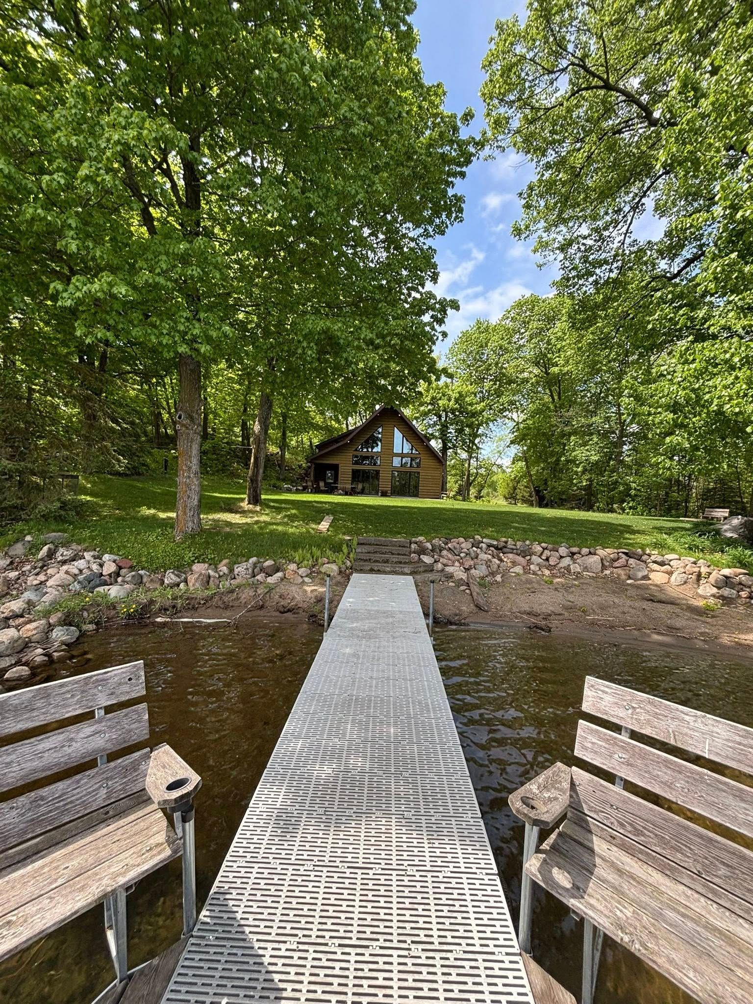 A metal dock extends from a lake toward a wooden house nestled among green trees under a bright blue sky.