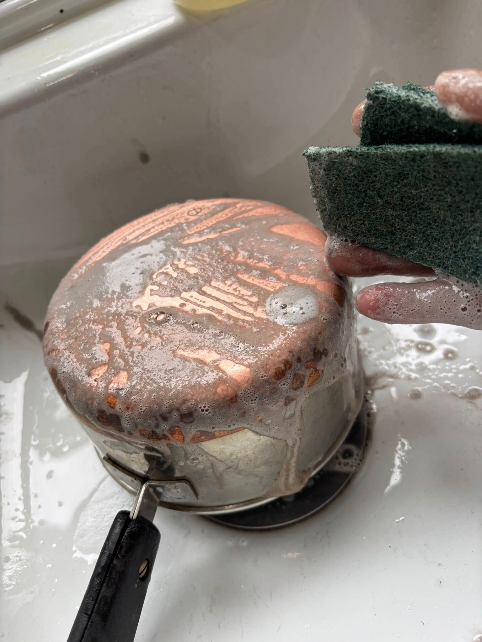 A person uses a green scouring pad to scrub soap suds off the copper-bottomed underside of a metal saucepan in a sink.