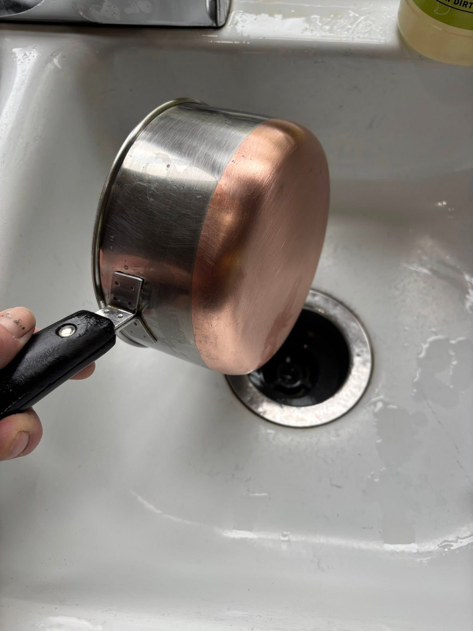 A hand holding a small stainless steel saucepan with a copper base over a white kitchen sink.
