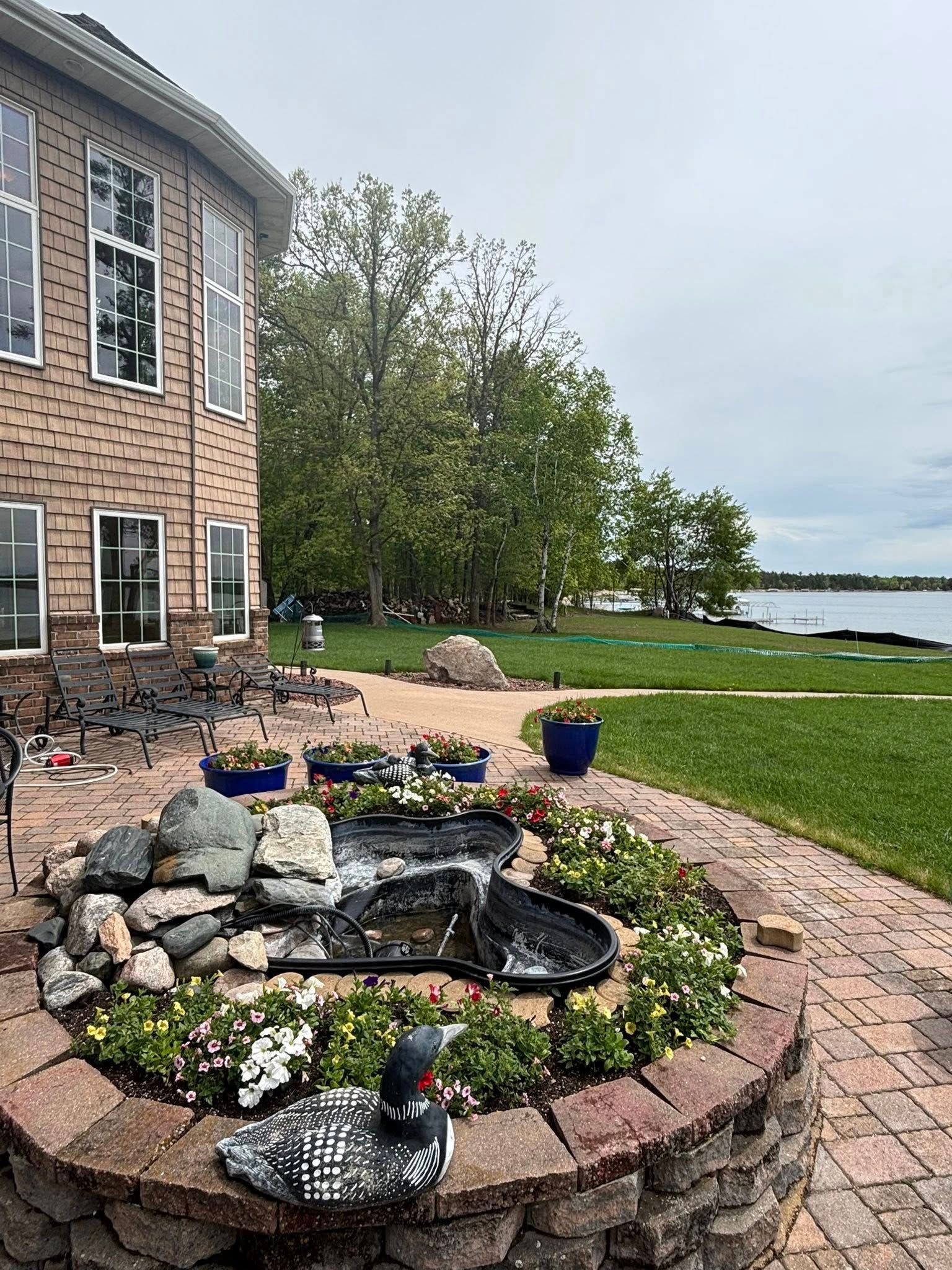 A patio with a stone fountain and a decorative loon figure, overlooking a grassy yard by a lake.