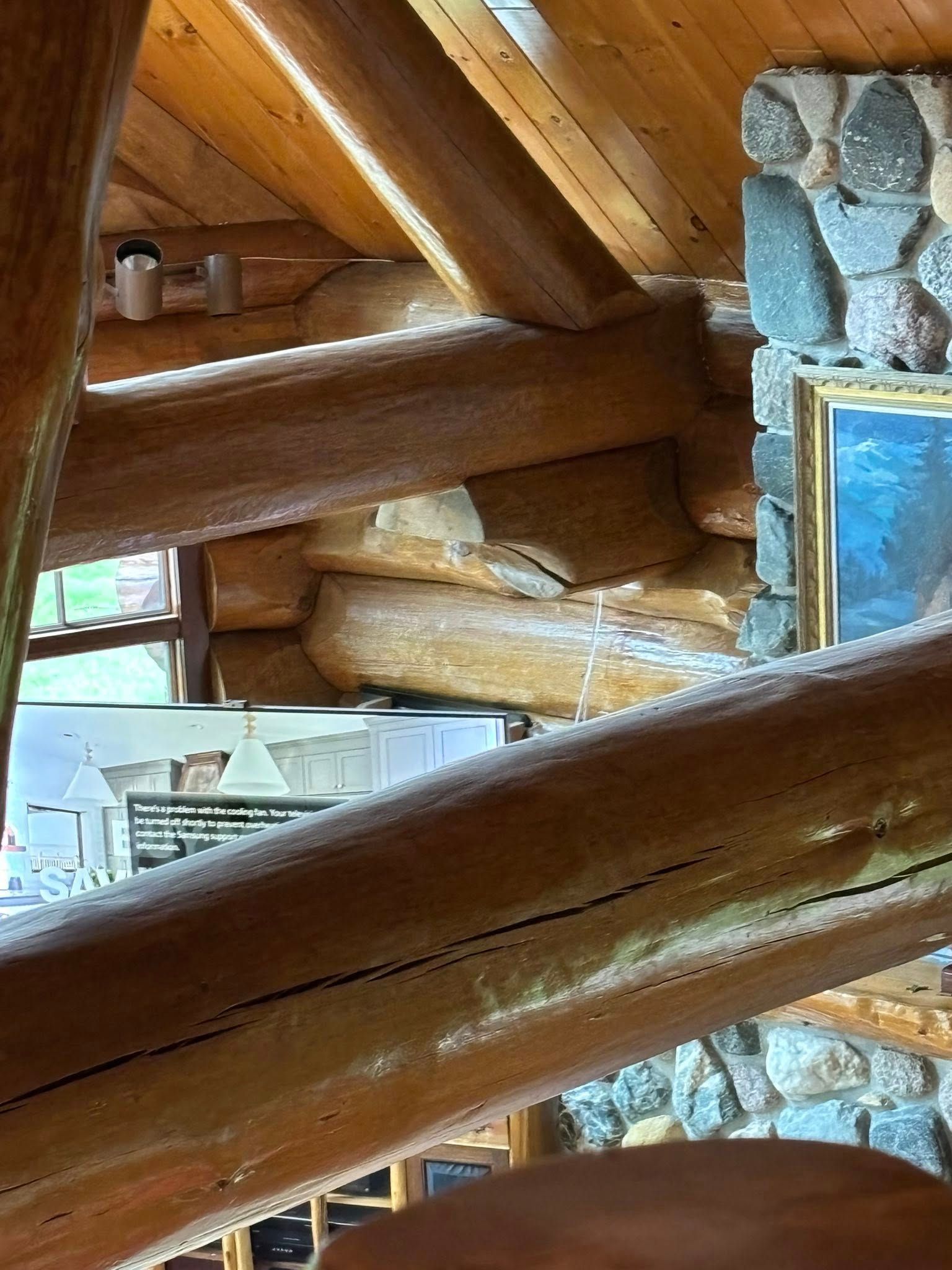 Wooden support beams in a rustic log cabin interior, with a stone fireplace and a framed picture in the background.