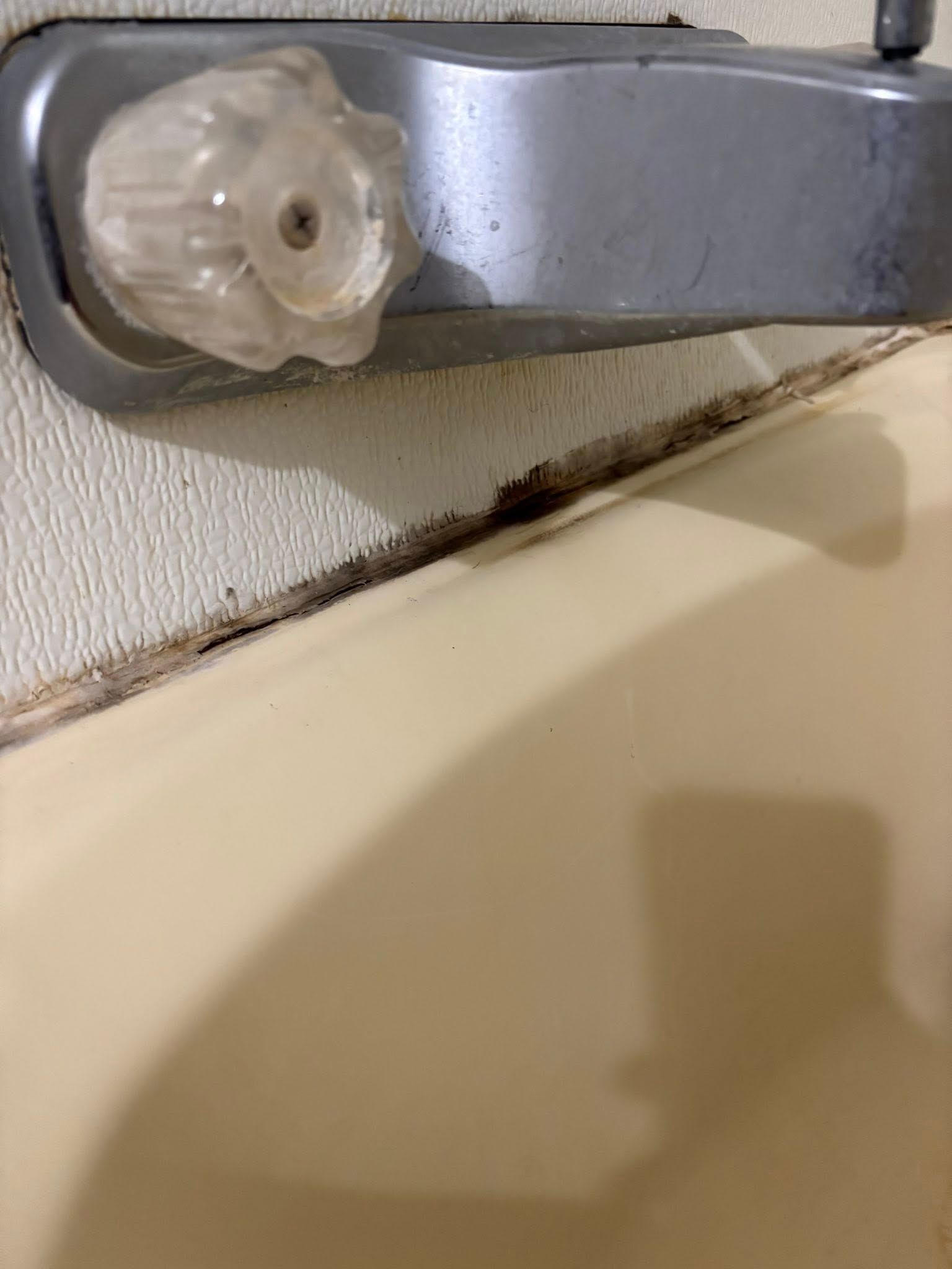 A close-up of a bathtub faucet handle with significant mold and damaged, cracked caulk along the wall-to-tub seam.