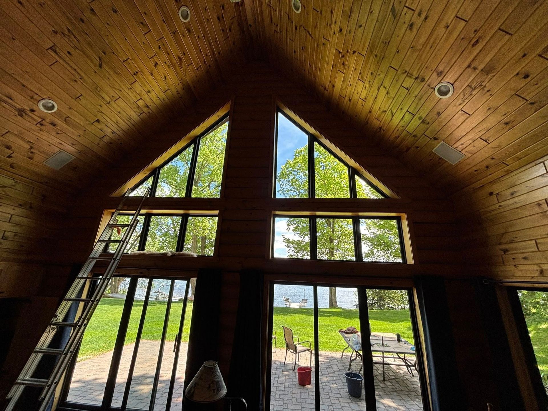 Interior view of a wooden cabin with large windows overlooking a lake. A ladder leans against the wall.