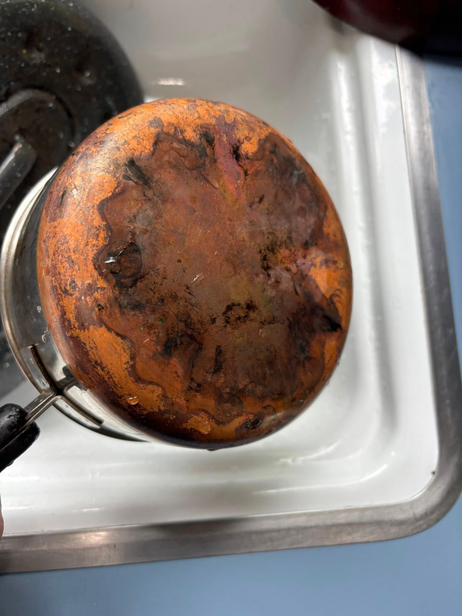 A close-up view of the rusted, discolored bottom of a metal pan held over a white kitchen sink.