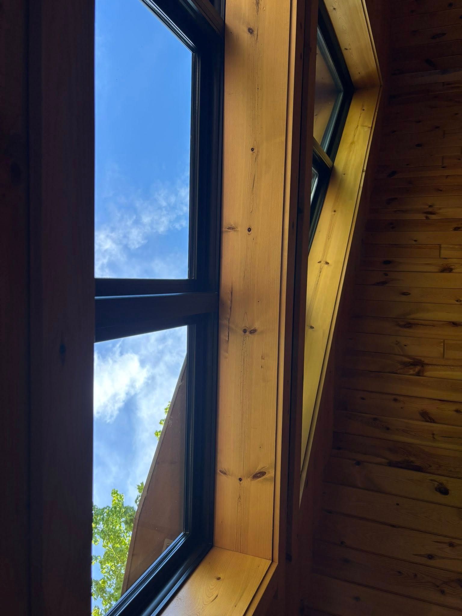 A low-angle view of a wooden-framed window looking out to a bright blue sky with clouds and green treetops.