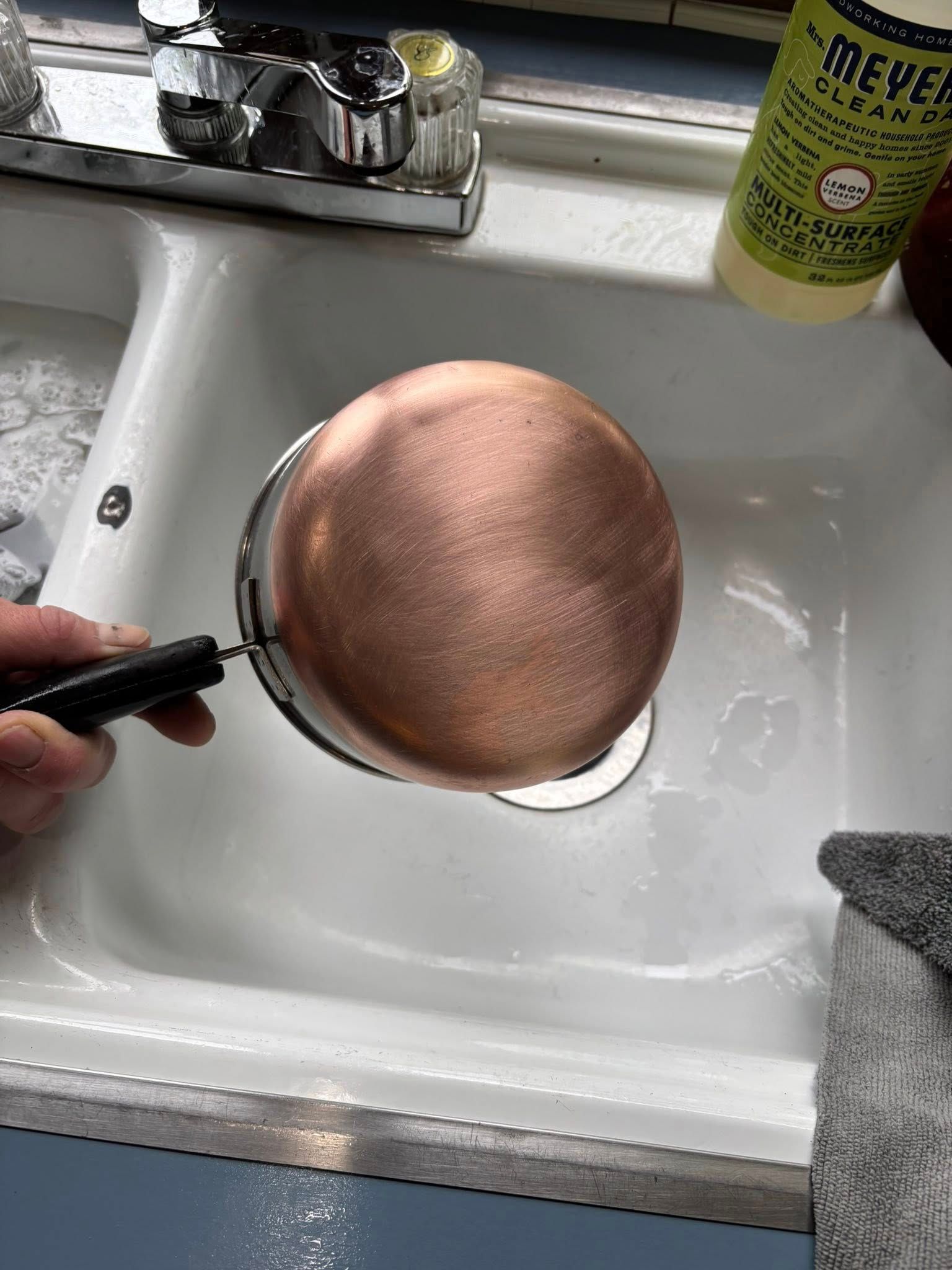 A person holds a copper-bottomed pot over a white kitchen sink.