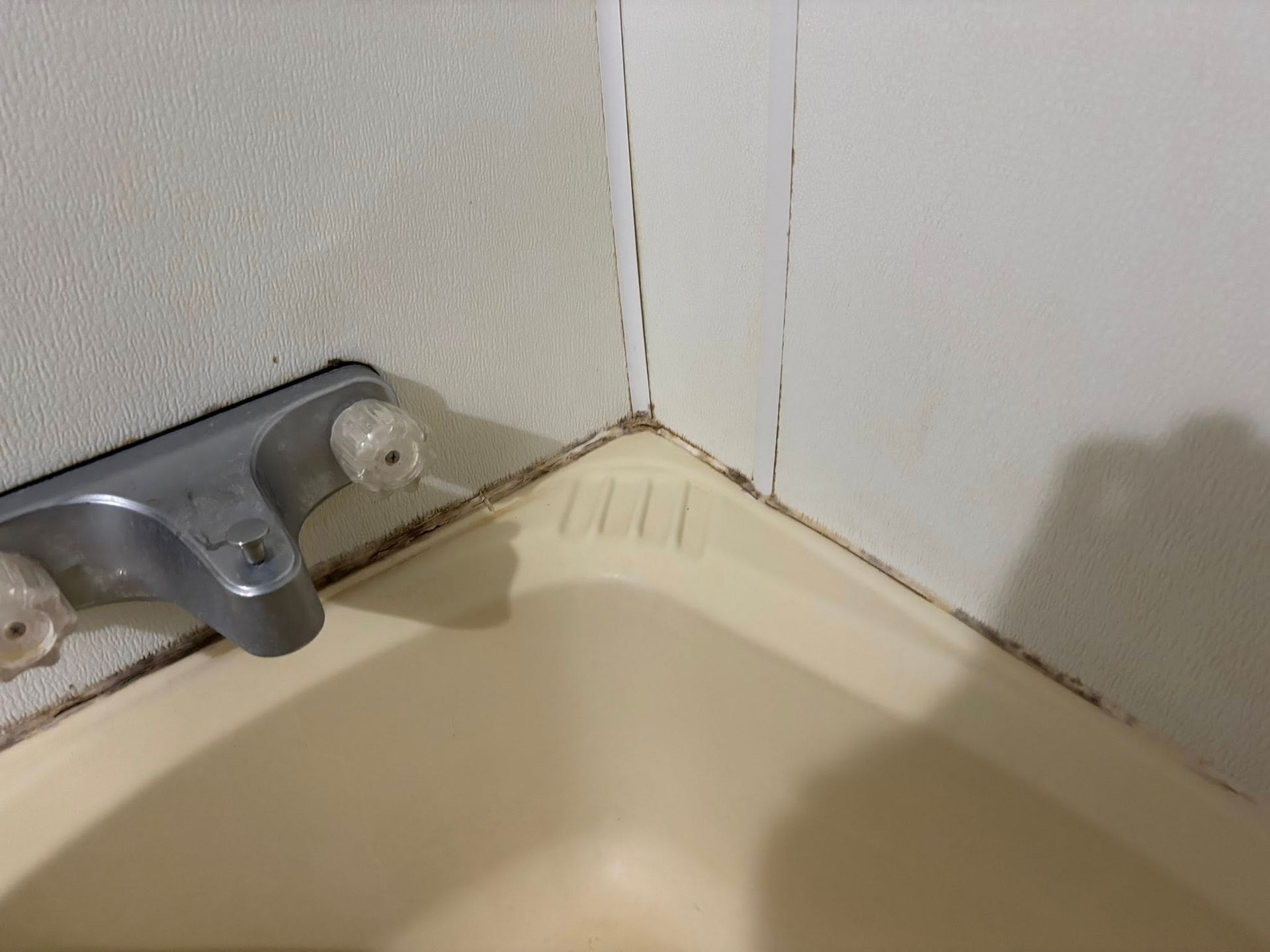 A close-up of a bathtub corner showing stained, cracked caulk along the wall and around the faucet assembly.