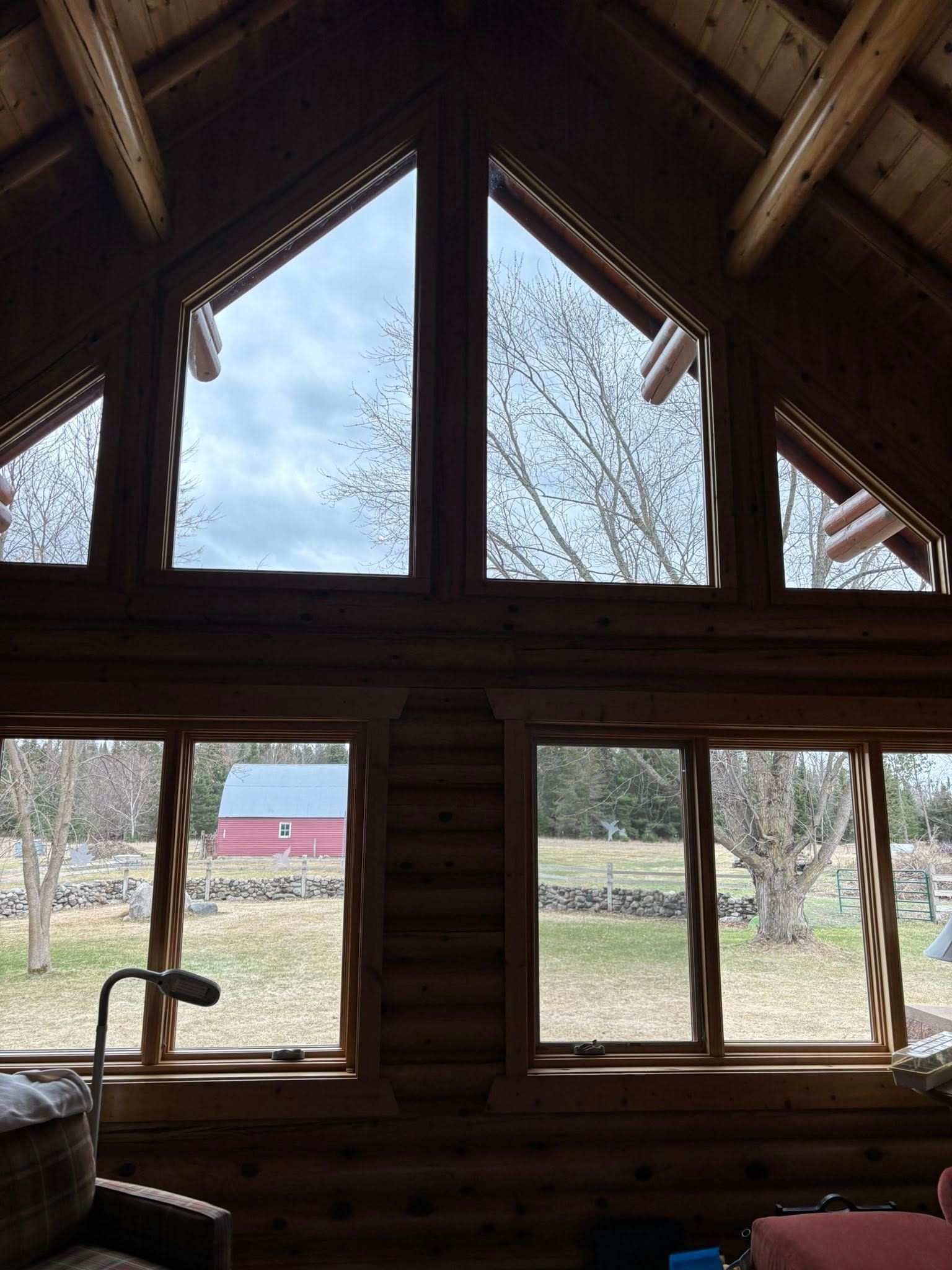 A view through the large, multi-pane windows of a rustic log cabin, looking out at a grassy field and a red barn.