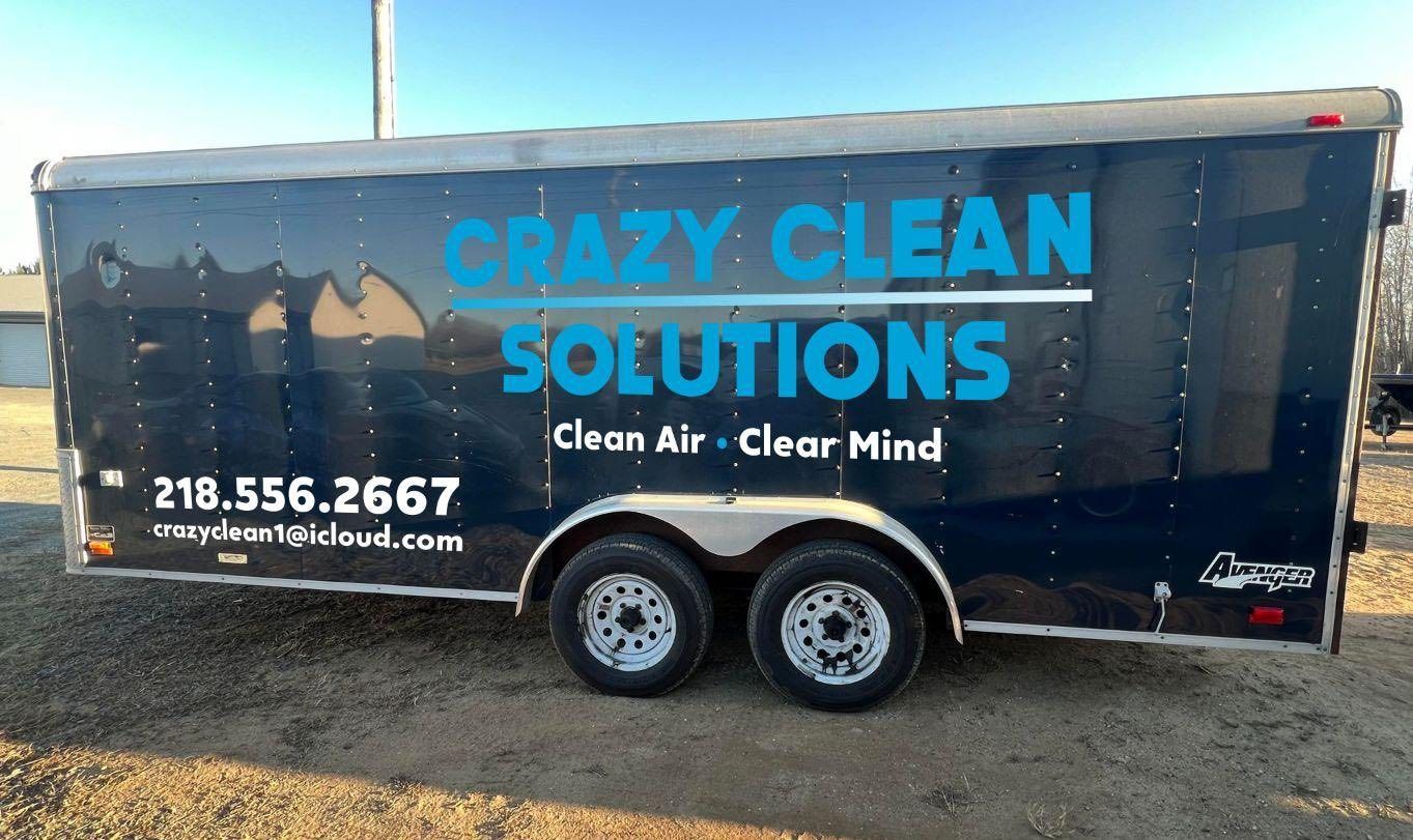 A black cargo trailer for Crazy Clean Solutions with blue text, a phone number, and email, parked on a gravel lot.