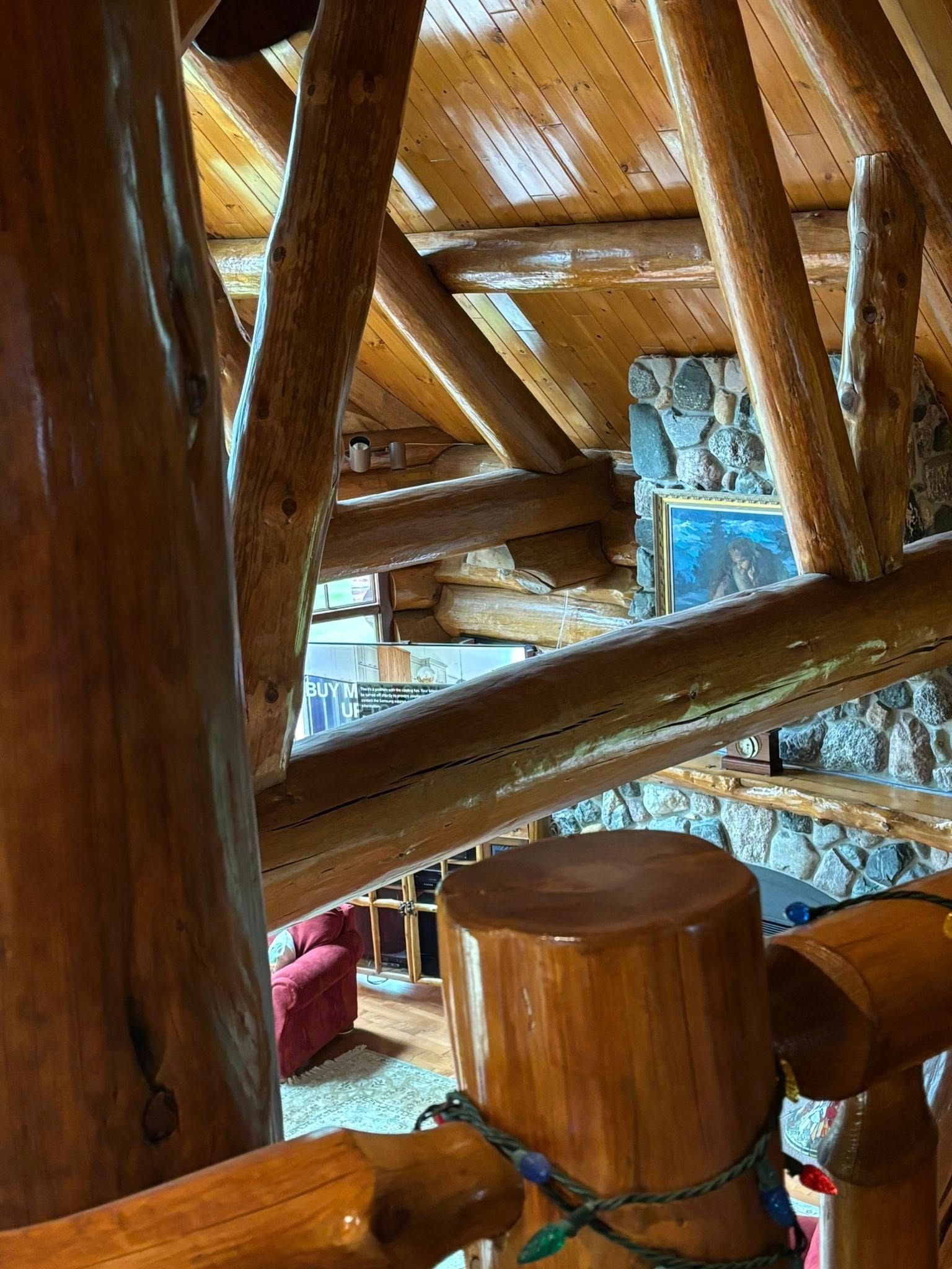 A high-angle view looking down through rustic wooden log beams toward a room with a stone fireplace and a red chair.