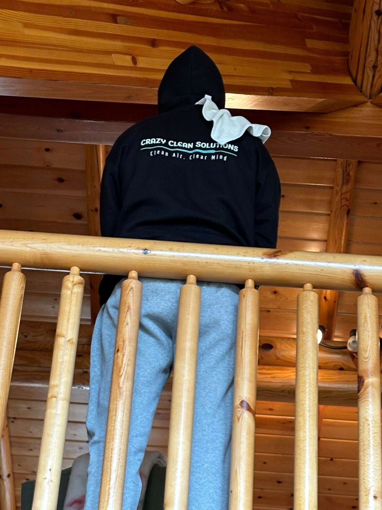 A person in a black hooded sweatshirt and gray sweatpants stands facing a wooden railing inside a log cabin.