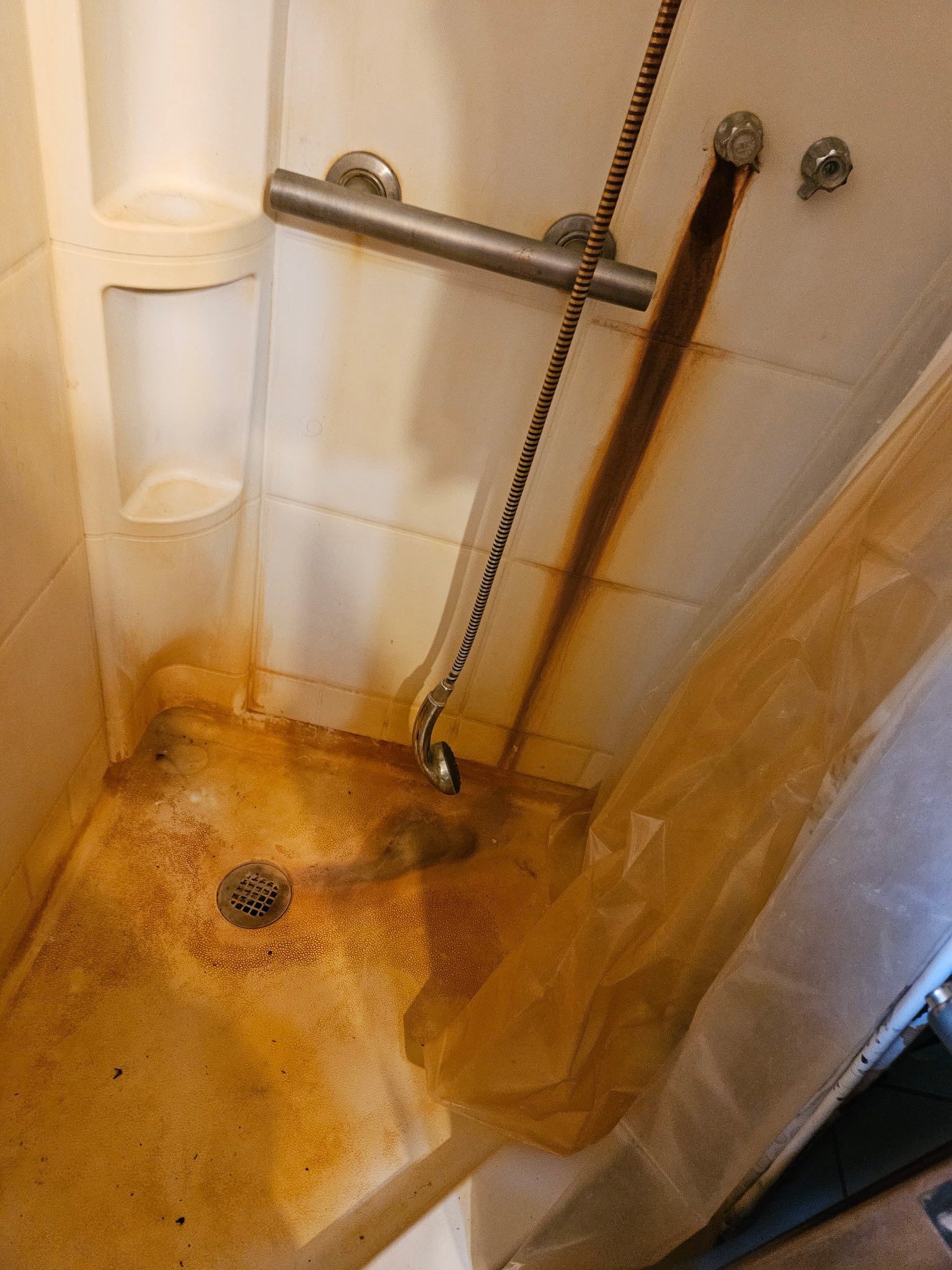 Shower stall featuring a metal grab bar and handheld showerhead, showing heavy orange-brown rust staining.