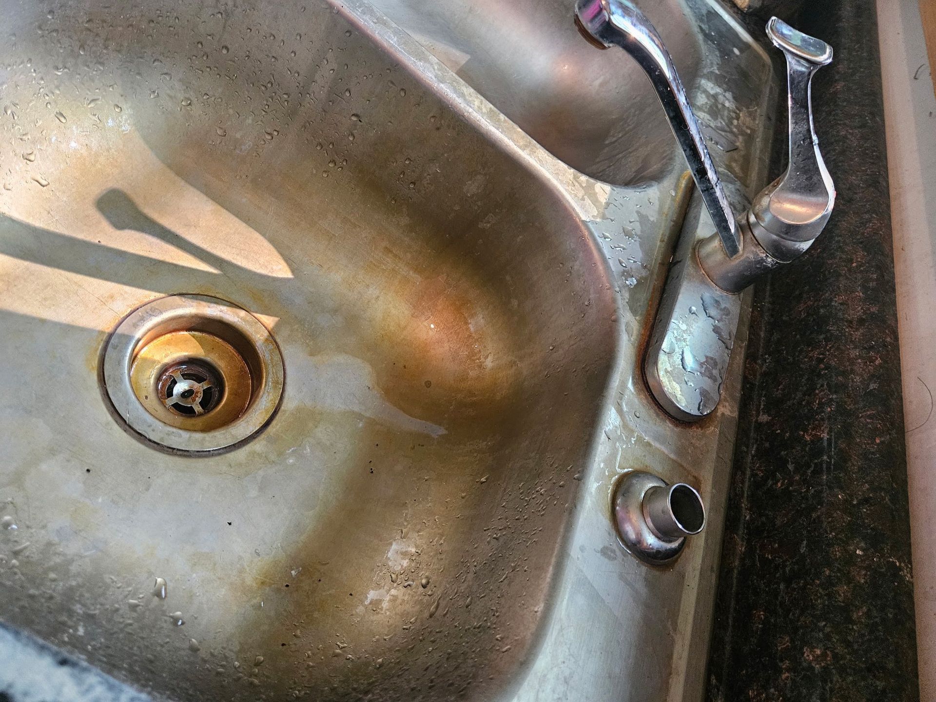 A stained stainless steel kitchen sink with a detached faucet handle and a missing faucet spout.