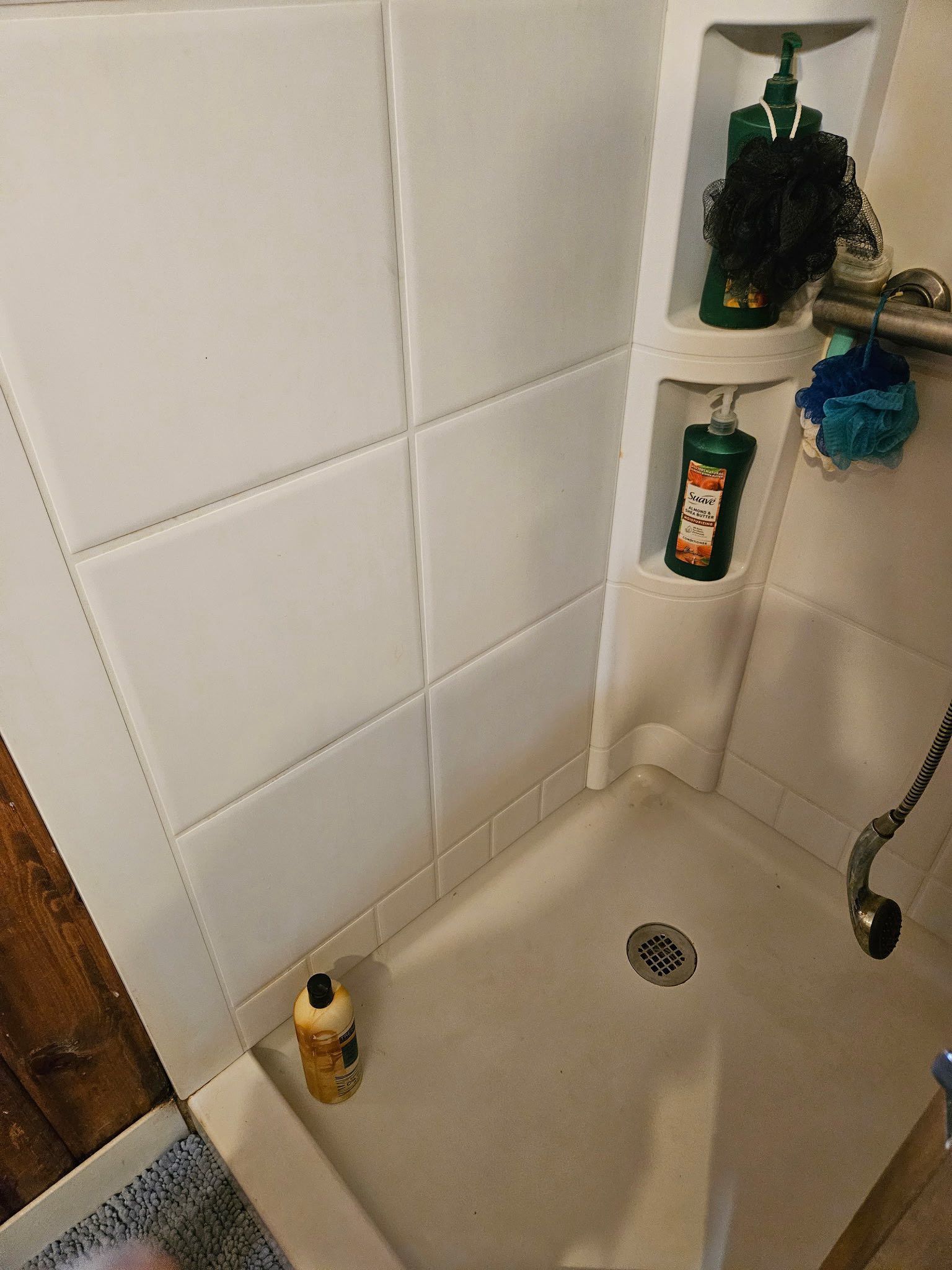 White shower stall featuring a corner shelving unit with bottles, sponges, and a drain on the floor.
