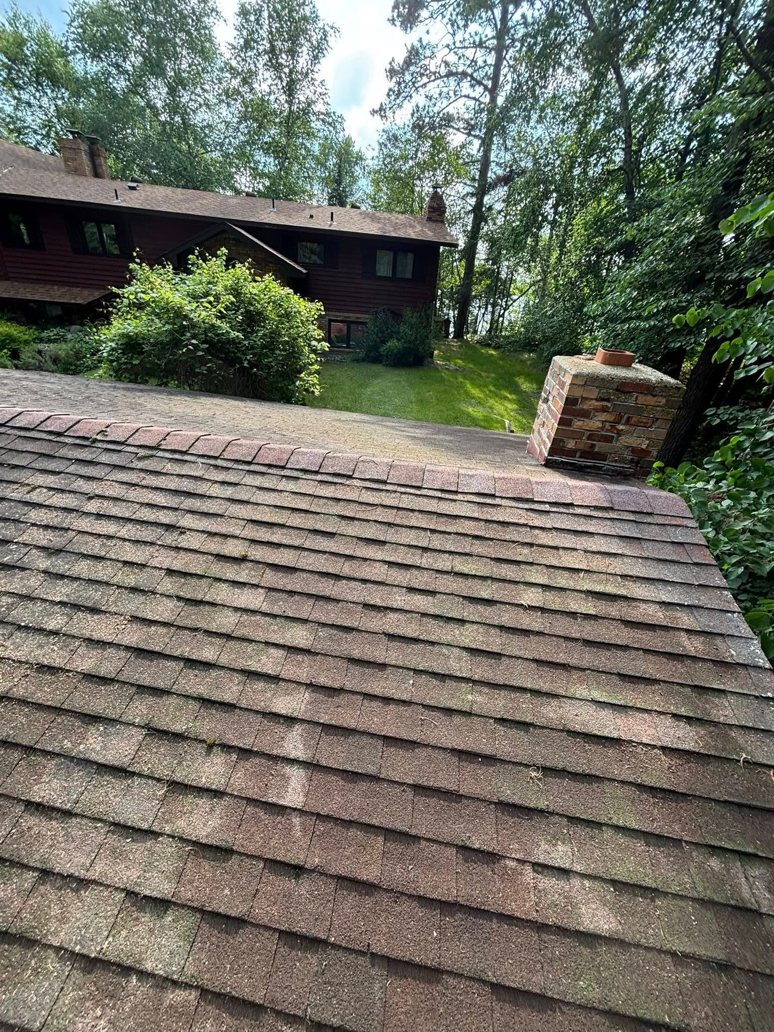 A view looking down over a weathered, brown shingled roof toward a wooded yard with a multi-story cabin and stone chimney.