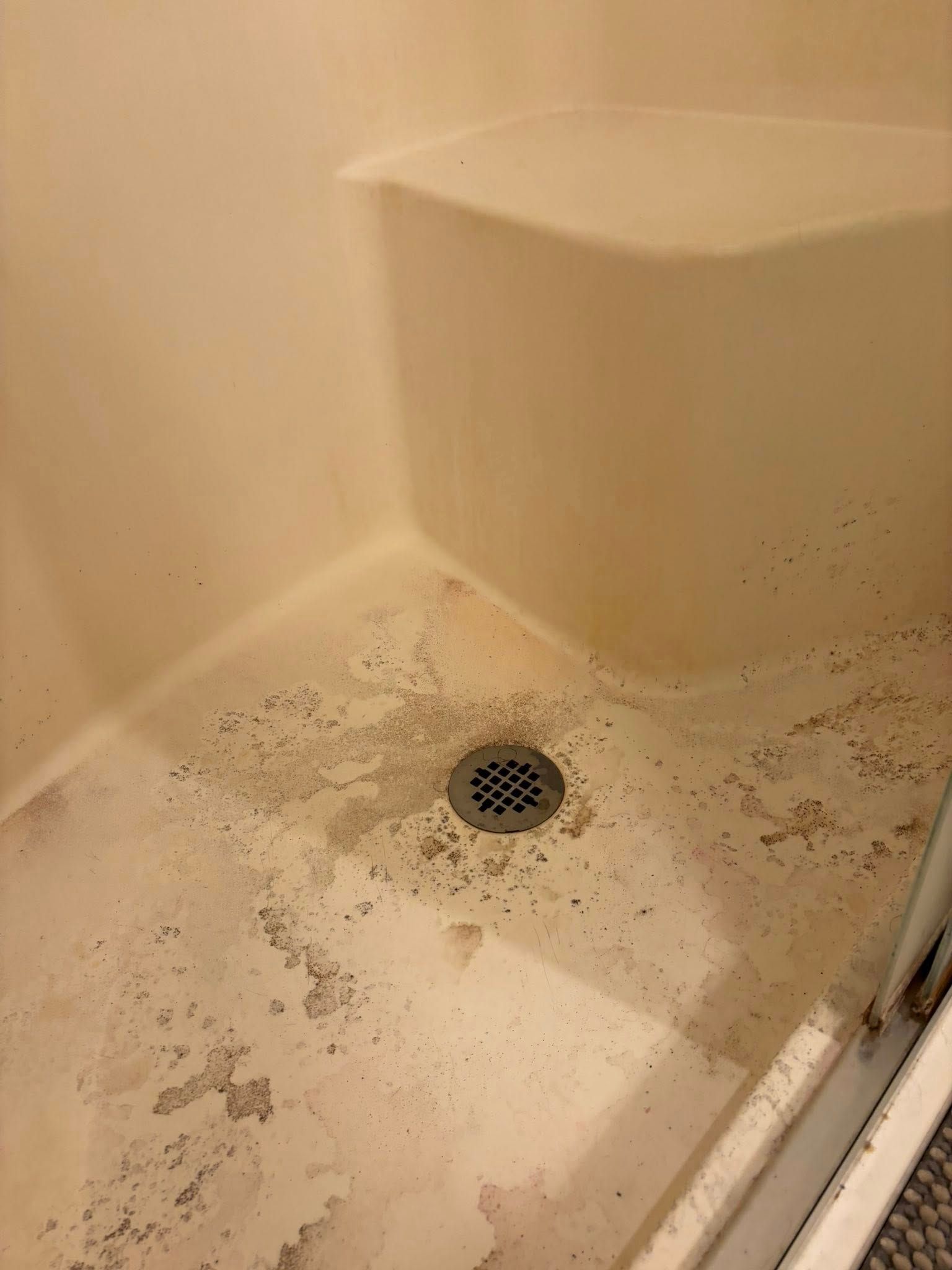 A shower stall with a built-in seat and a drain, featuring significant staining and damaged, peeling floor finish.