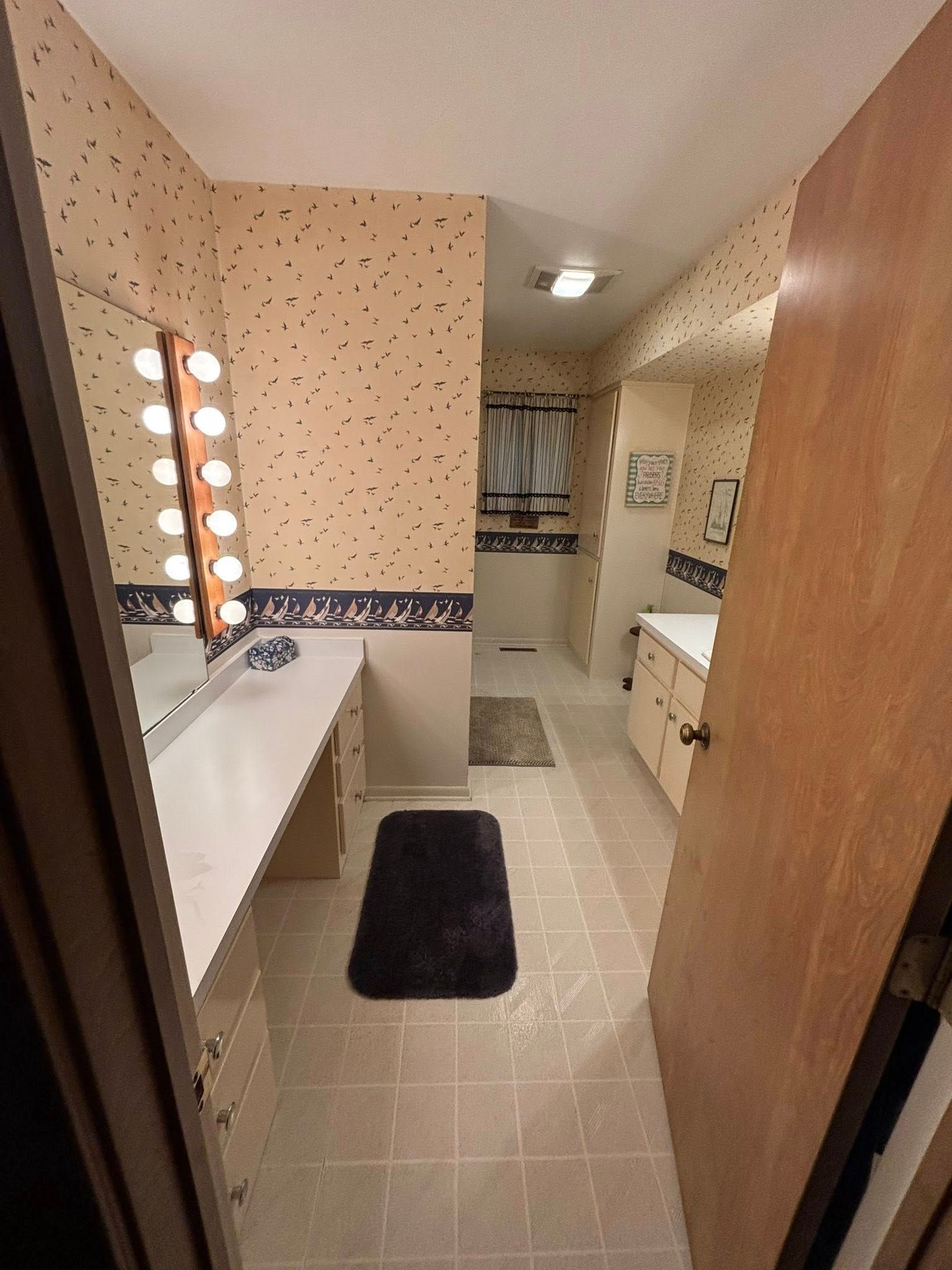A bathroom vanity with hollywood-style lights, patterned wallpaper, a white countertop, and tiled floors.