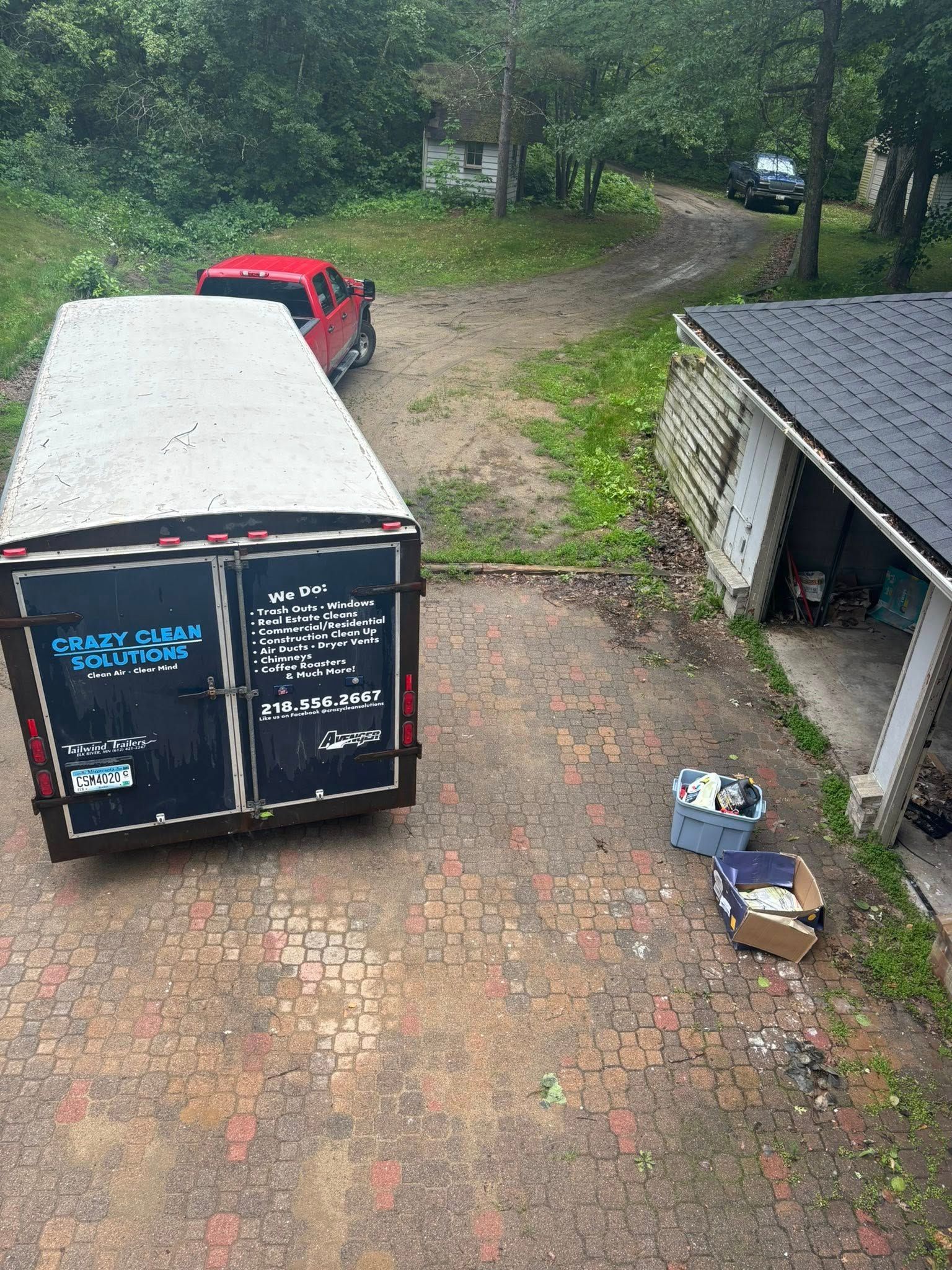 A red pickup truck is parked in a tree-lined driveway next to a large utility trailer and a small building.