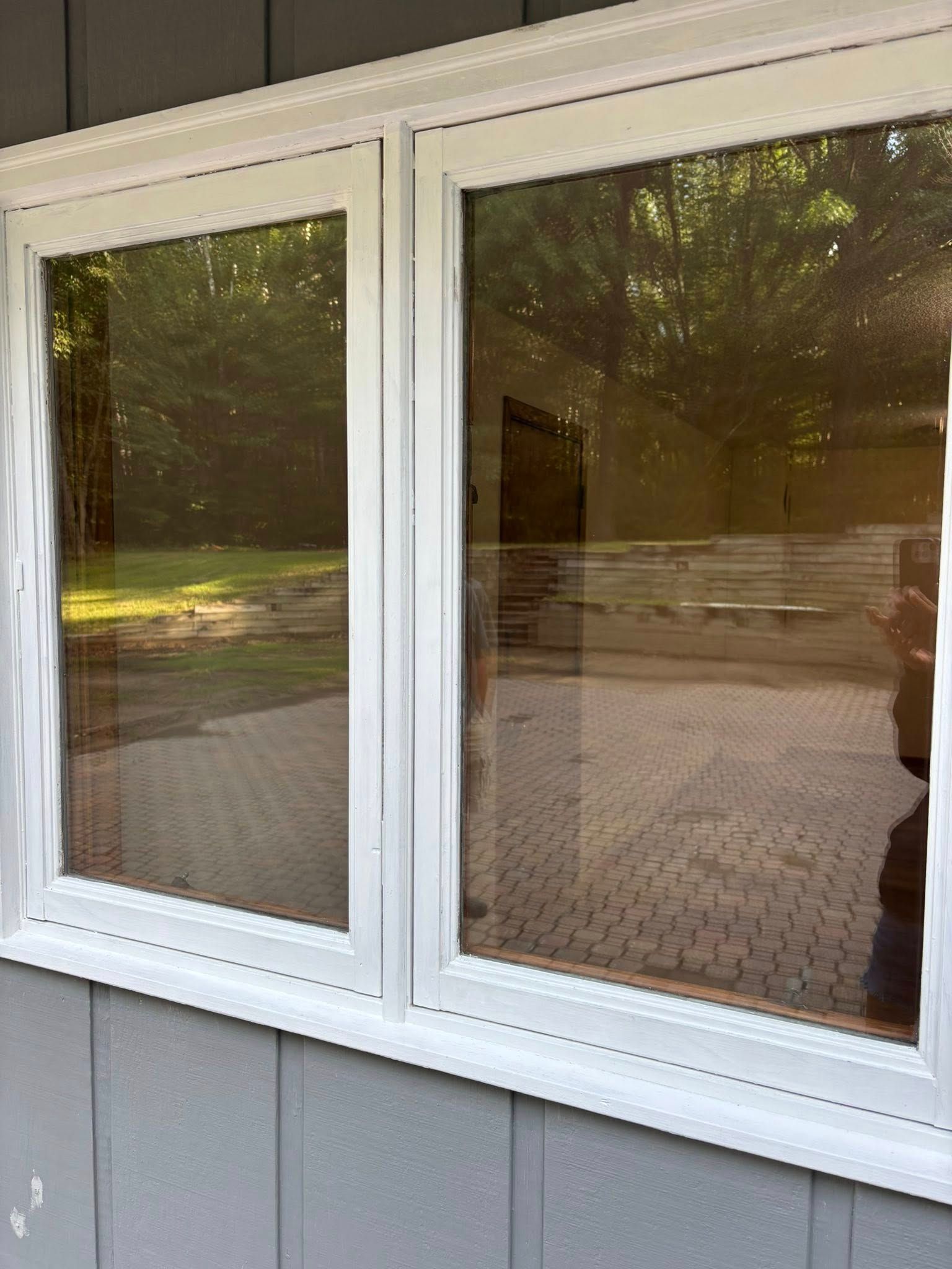 White-framed windows reflect a brick patio and trees outside, set against gray siding.
