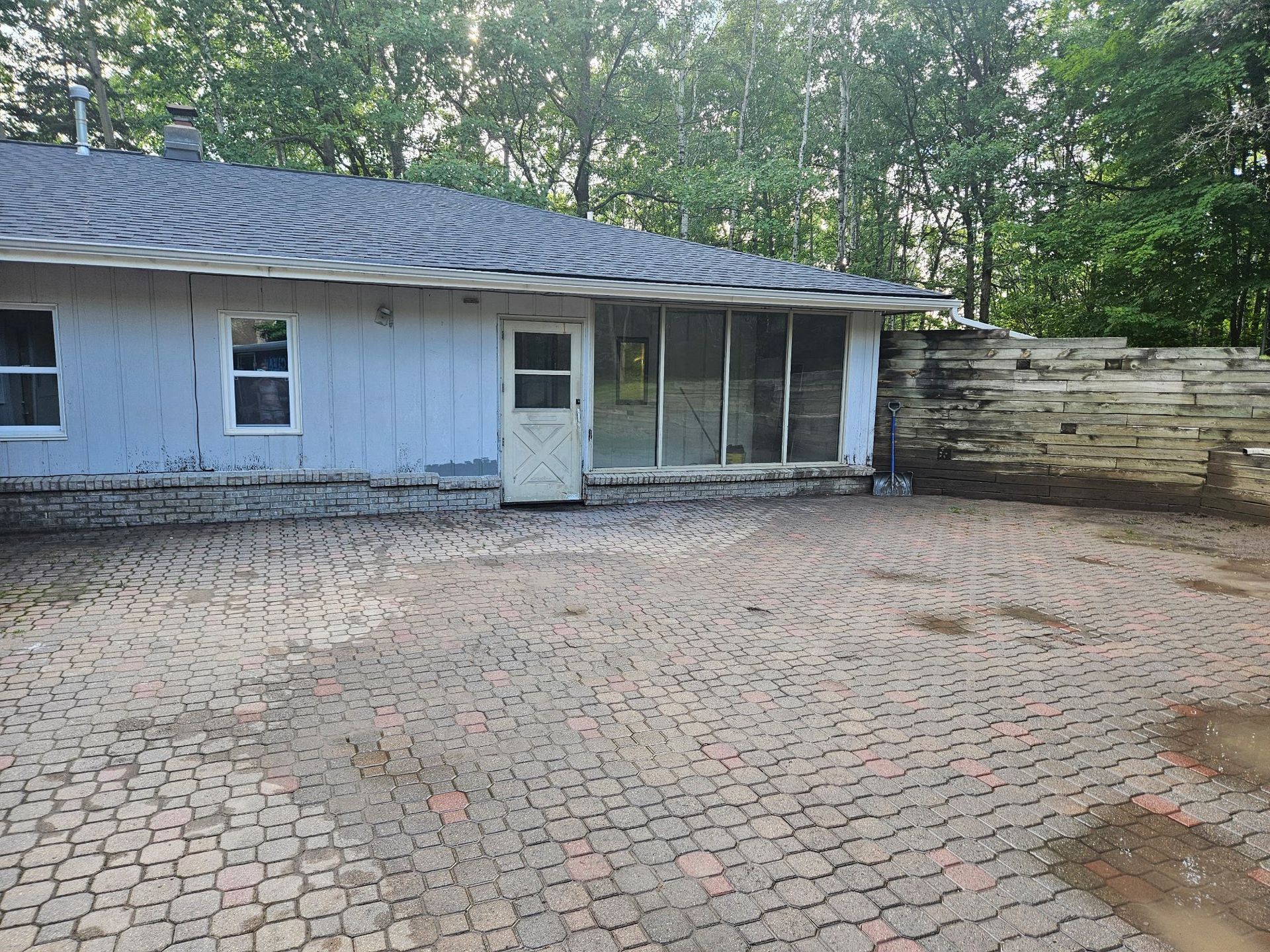 A light-colored house with a patio made of interlocking pavers, leading to a wooden fence and surrounding trees.
