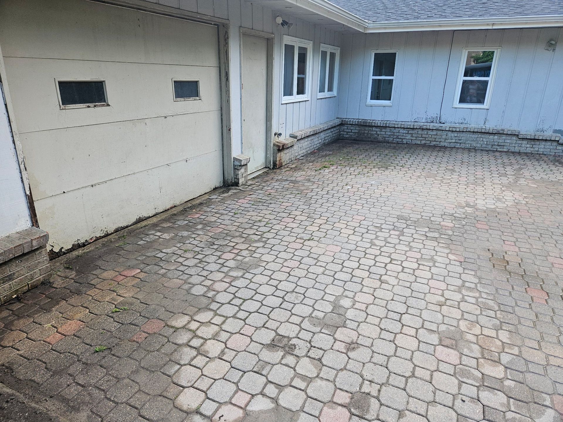 A paved stone driveway leads up to a garage door and house exterior with blue siding and white-framed windows.