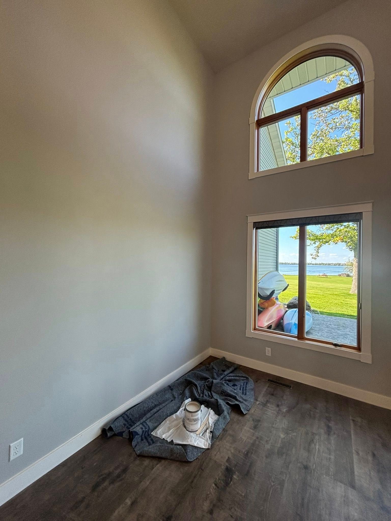 A room with gray walls, dark wood floors, and two stacked windows overlooking a grassy yard and water.