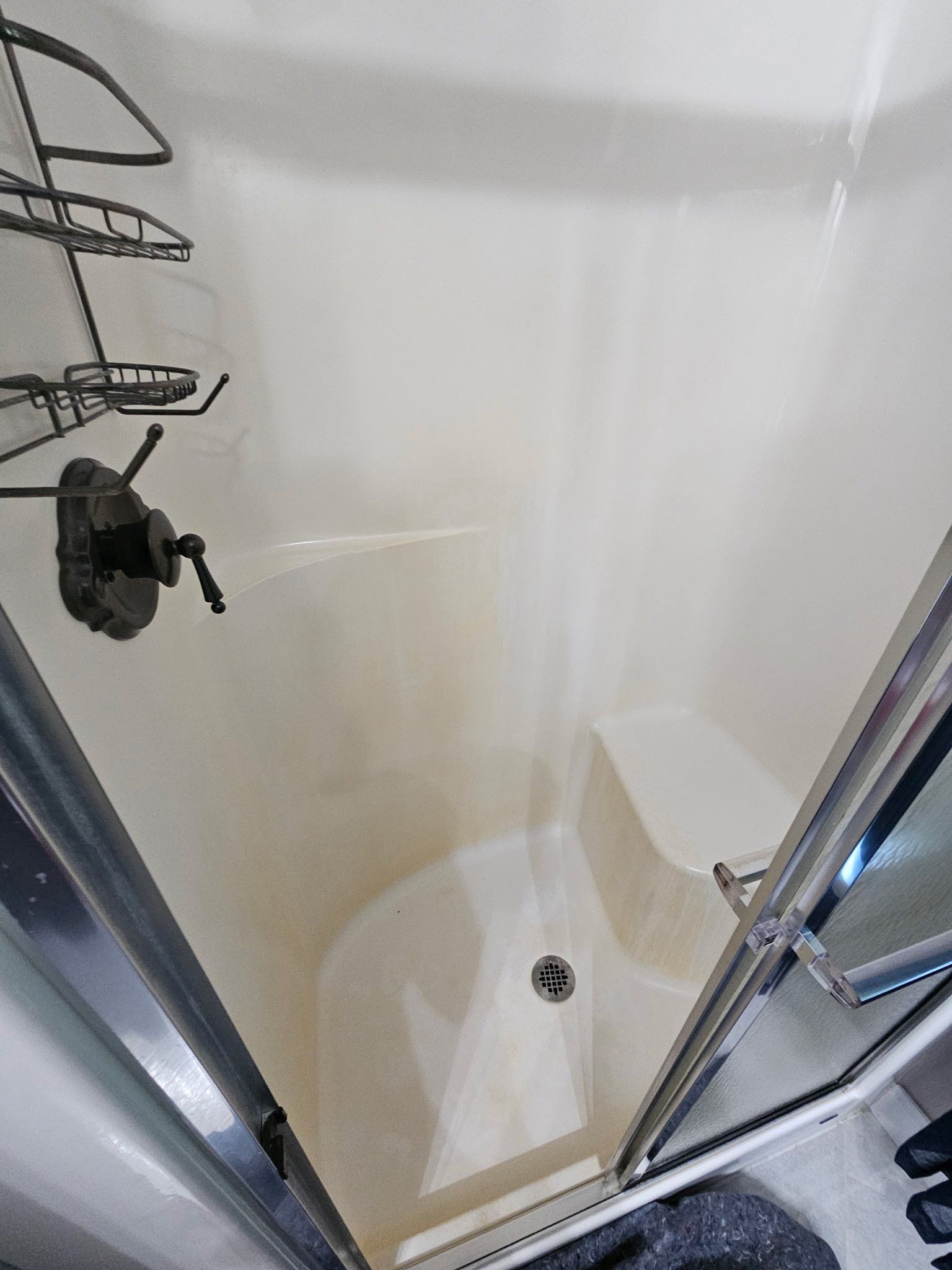 A white, walk-in shower stall with an attached wire storage rack, a black faucet handle, and a built-in corner seat.
