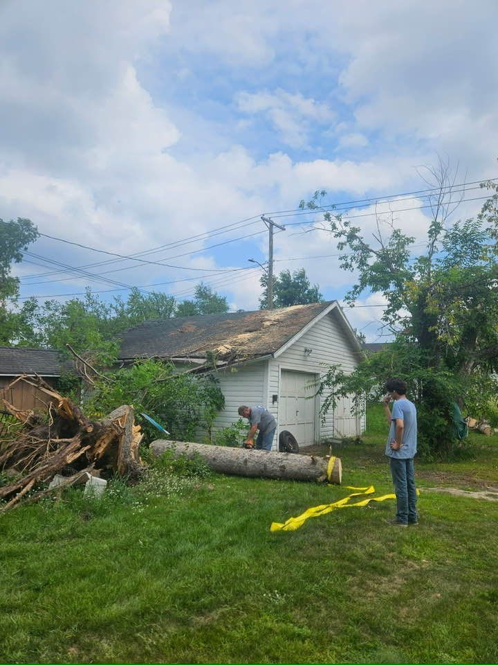 Two people working on removing a fallen tree from the roof of a detached garage on a grassy lot.