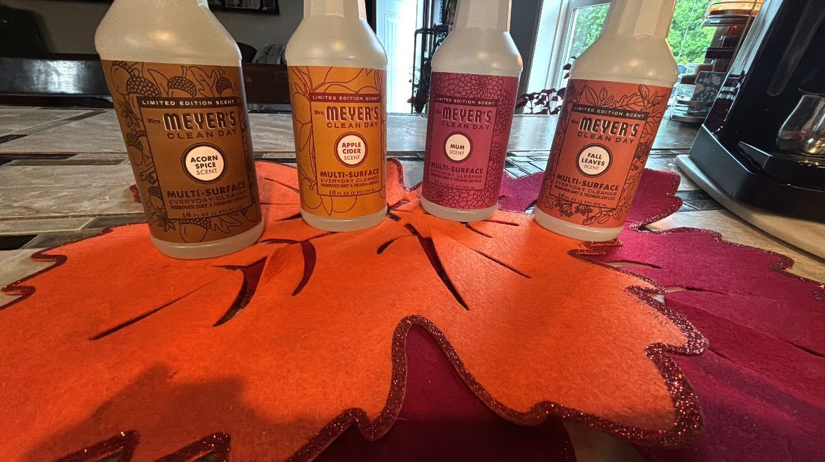 Four spray bottles with decorative fall-themed labels arranged on a felt leaf placemat on a wooden table.
