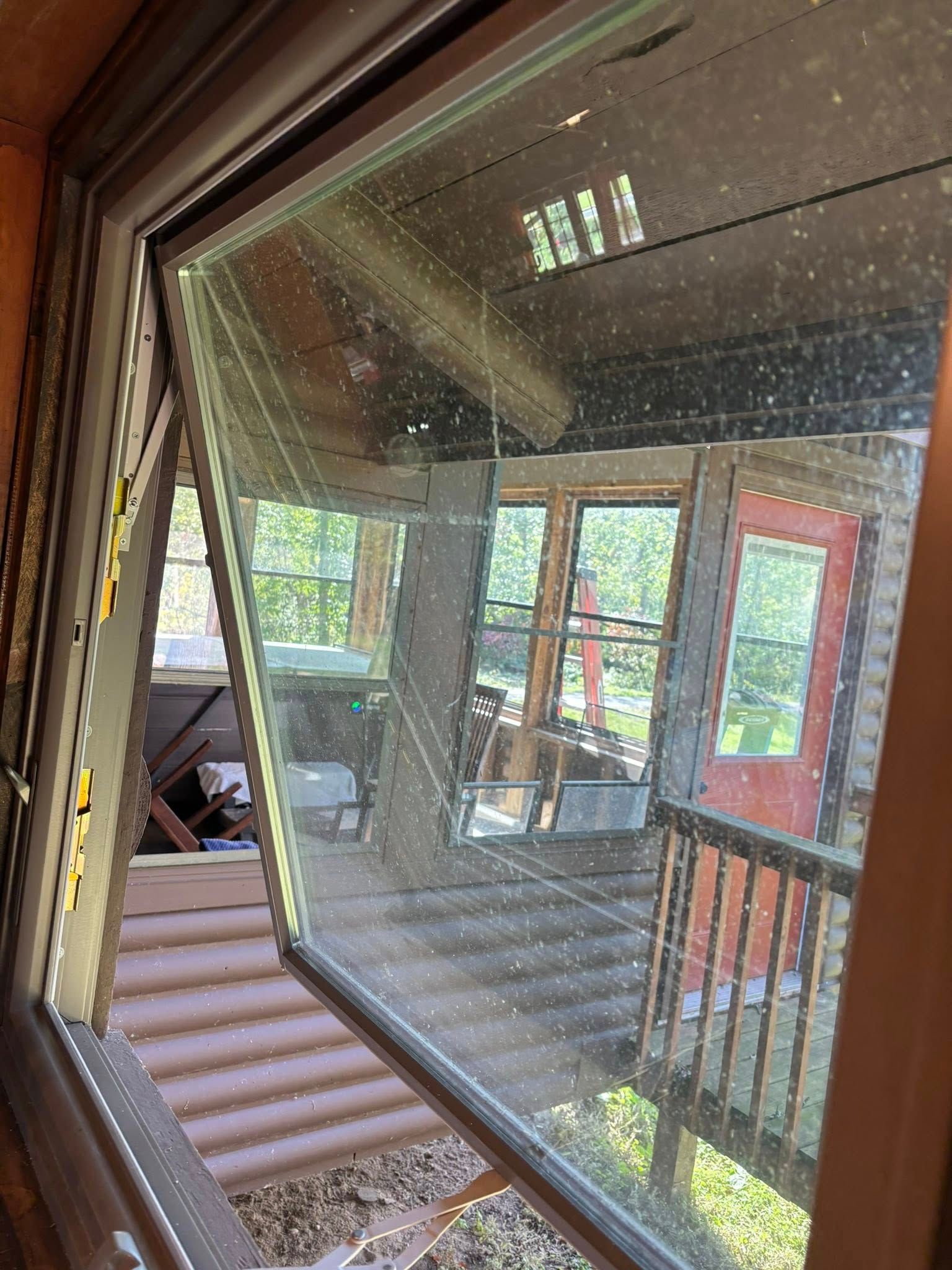 An open, outward-tilting casement window showing a reflected view of a porch and trees.
