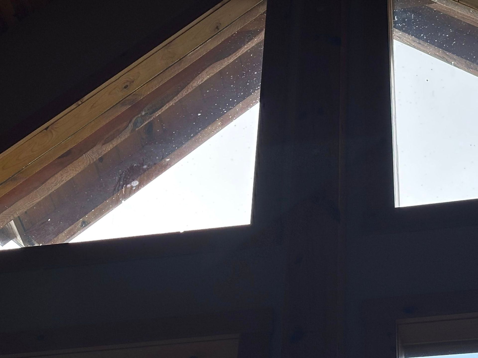 A view from inside looking out through a window at a cloudy sky, framed by dark interior trim and a wooden roof beam.