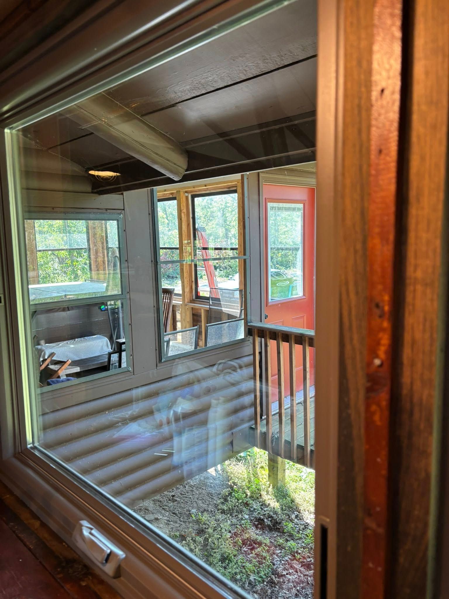A view through a window onto a rustic wooden cabin porch, showing exterior walls, a railing, and surrounding greenery.