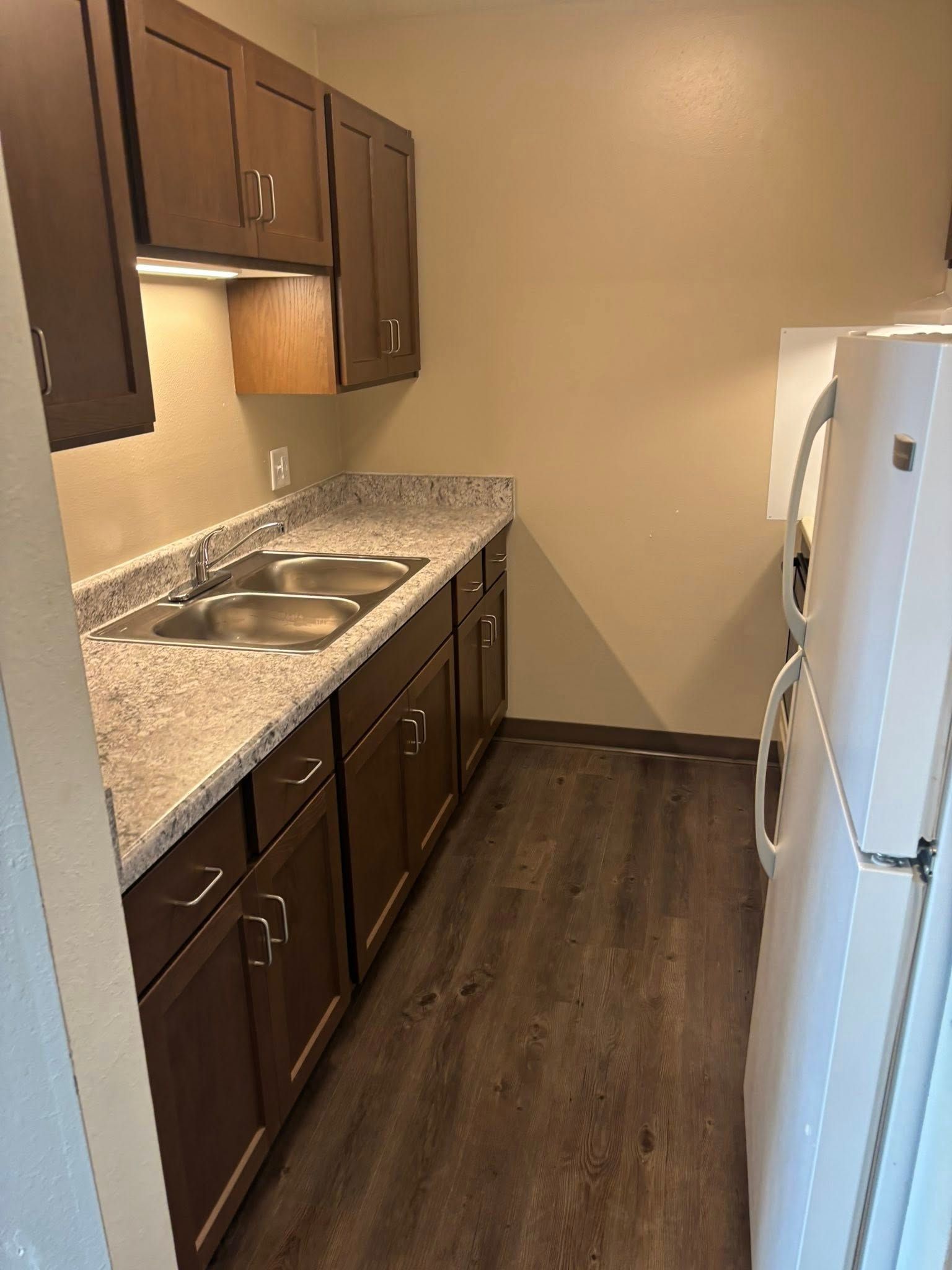 Galley kitchen with dark wood cabinets, light granite countertops, a stainless steel sink, and a white refrigerator.