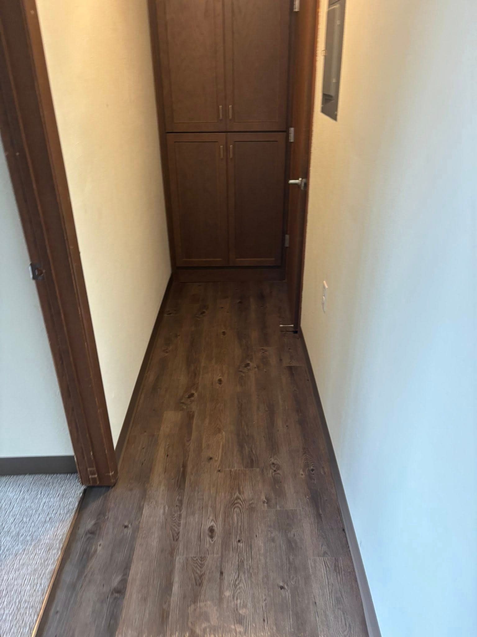 A narrow hallway with dark wood-look vinyl flooring, beige walls, and a tall, dark wooden built-in cabinet at the end.