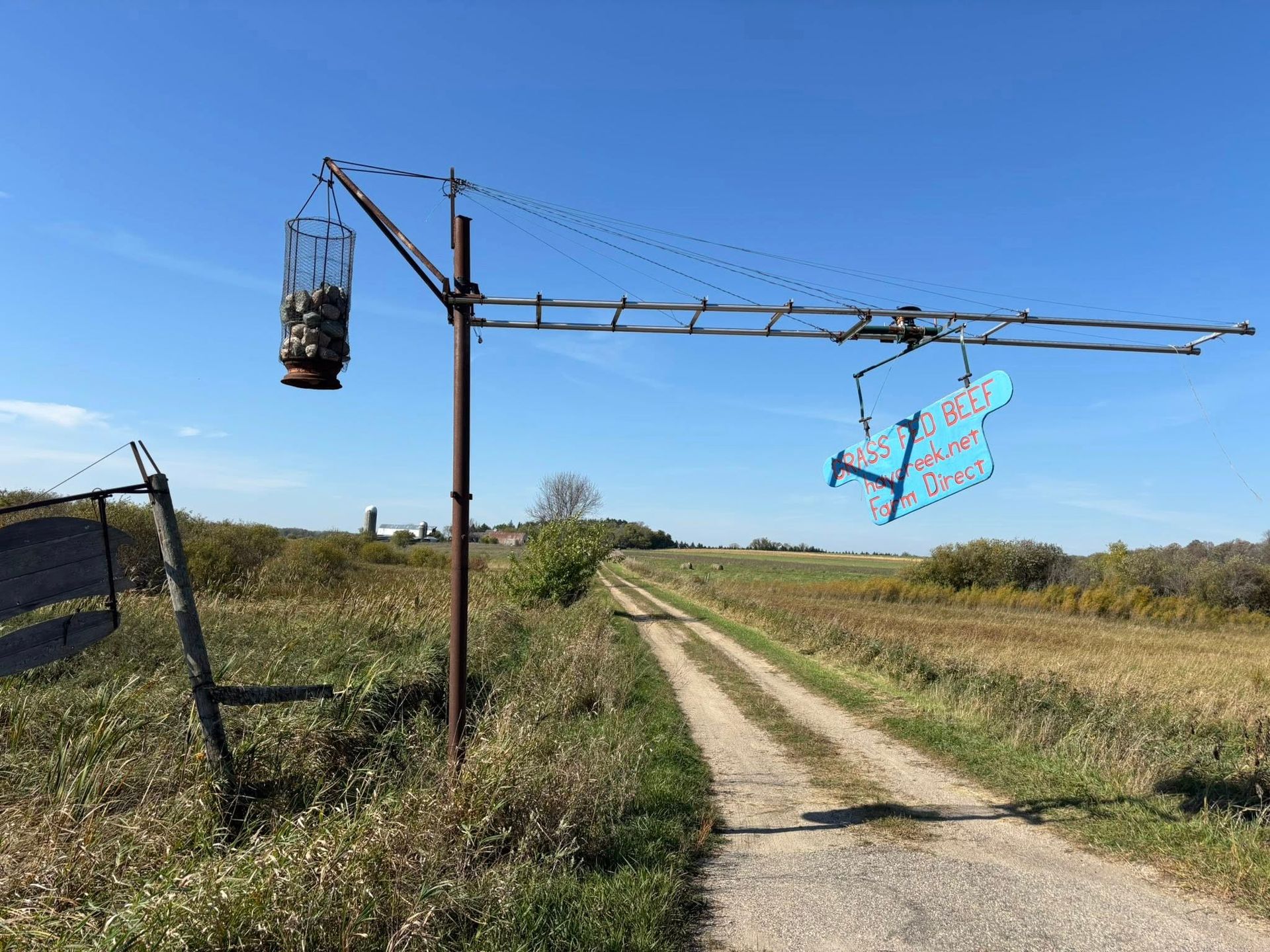 A metal structure over a dirt road holds a cage of rocks on one side and a blue, airplane-shaped sign on the other.
